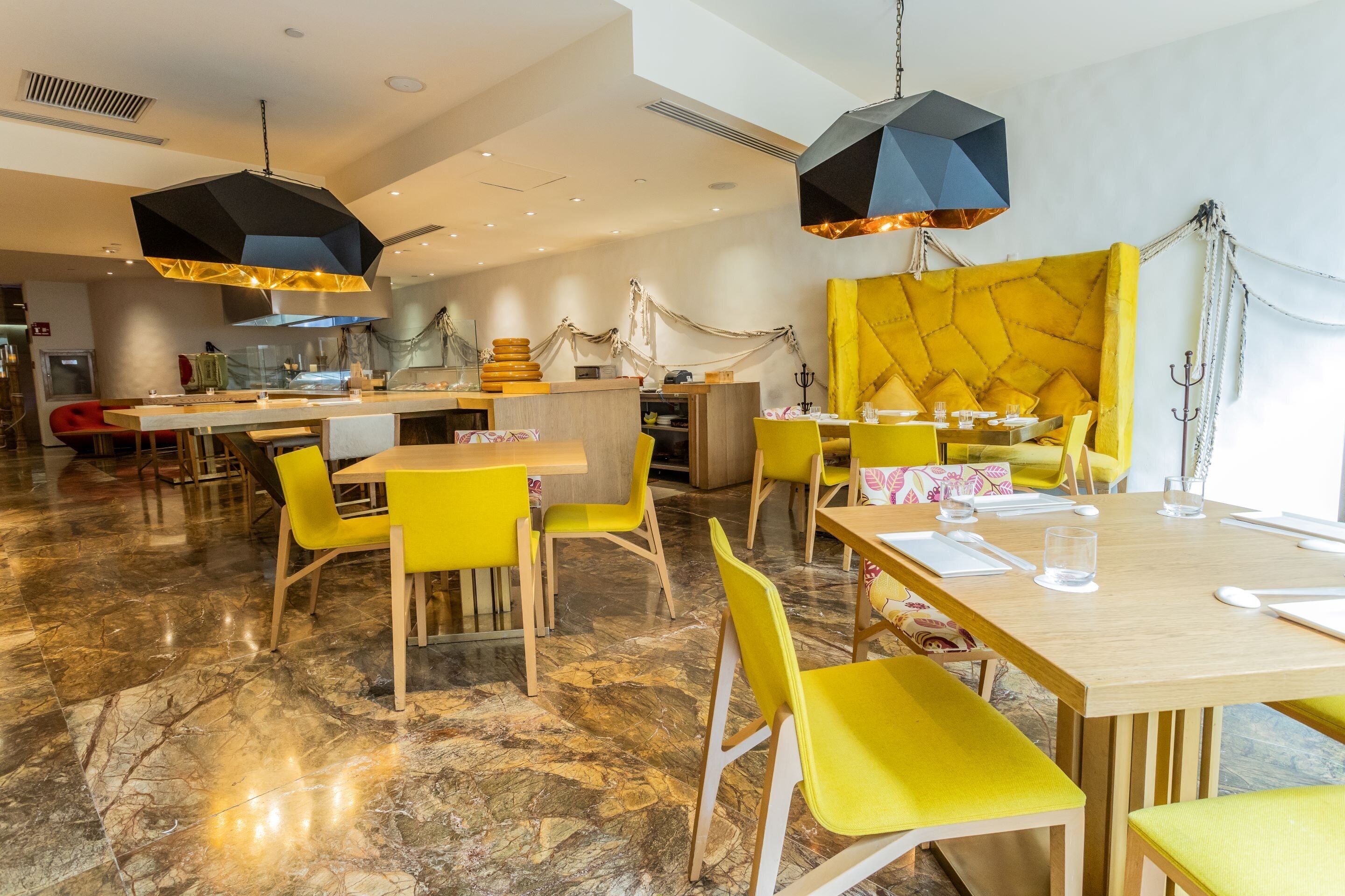 Un restaurante moderno con sillas y mesas amarillas, suelo de mármol e iluminación colgante geométrica.