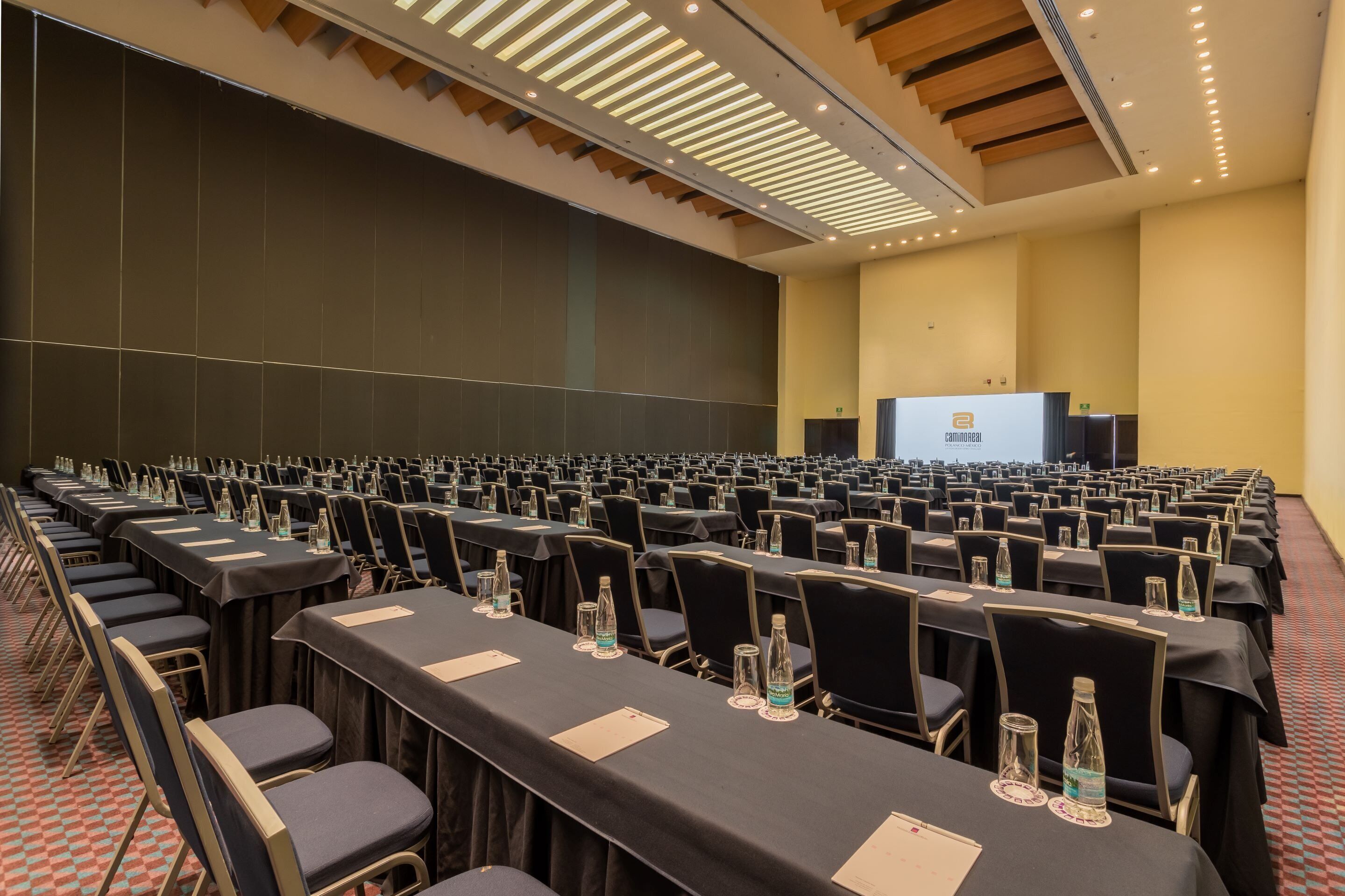 A conference room with rows of tables and chairs, a stage with a presentation screen, and a high ceiling with recessed lighting.