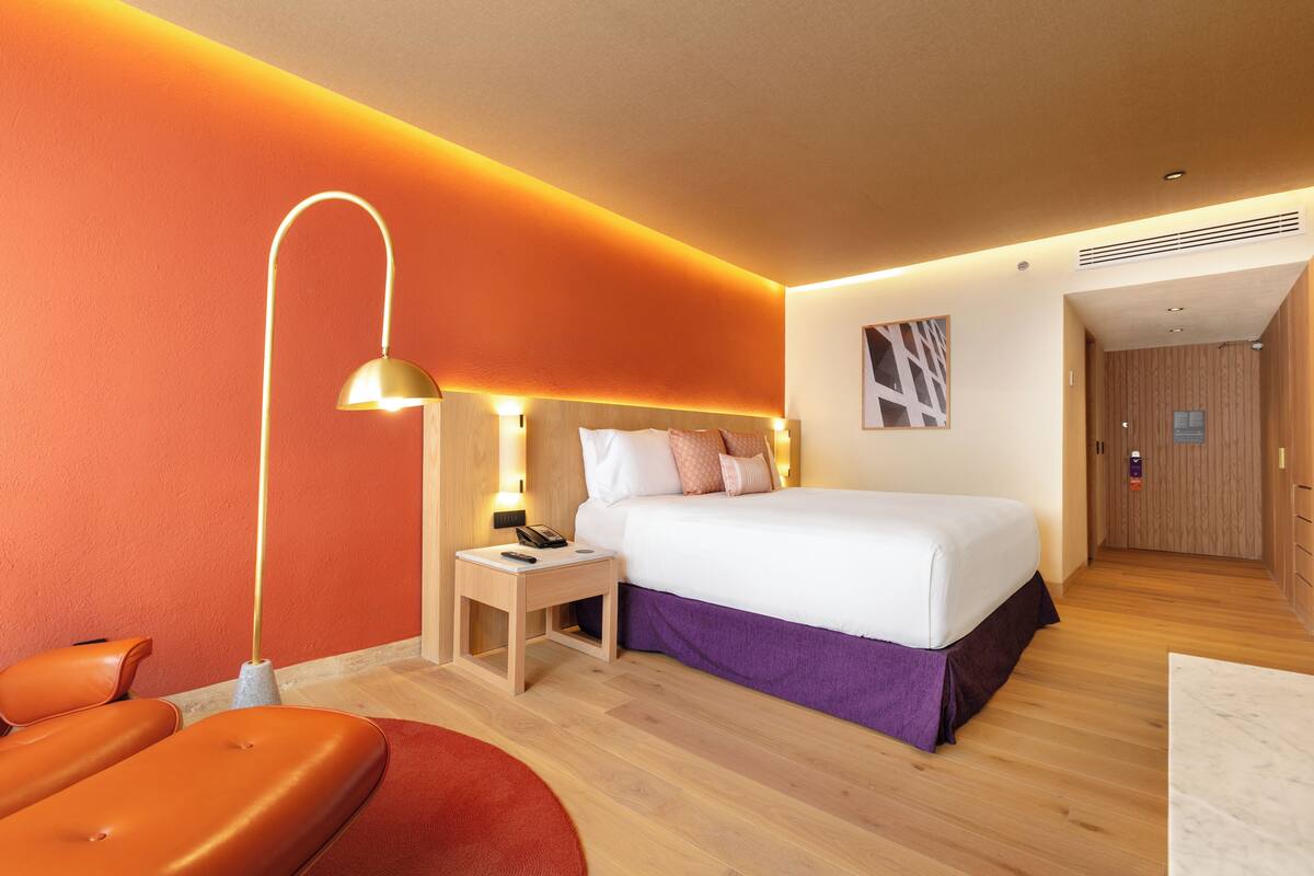 A modern hotel room with a large bed, a nightstand with a lamp, a wooden floor, and a red accent wall.
