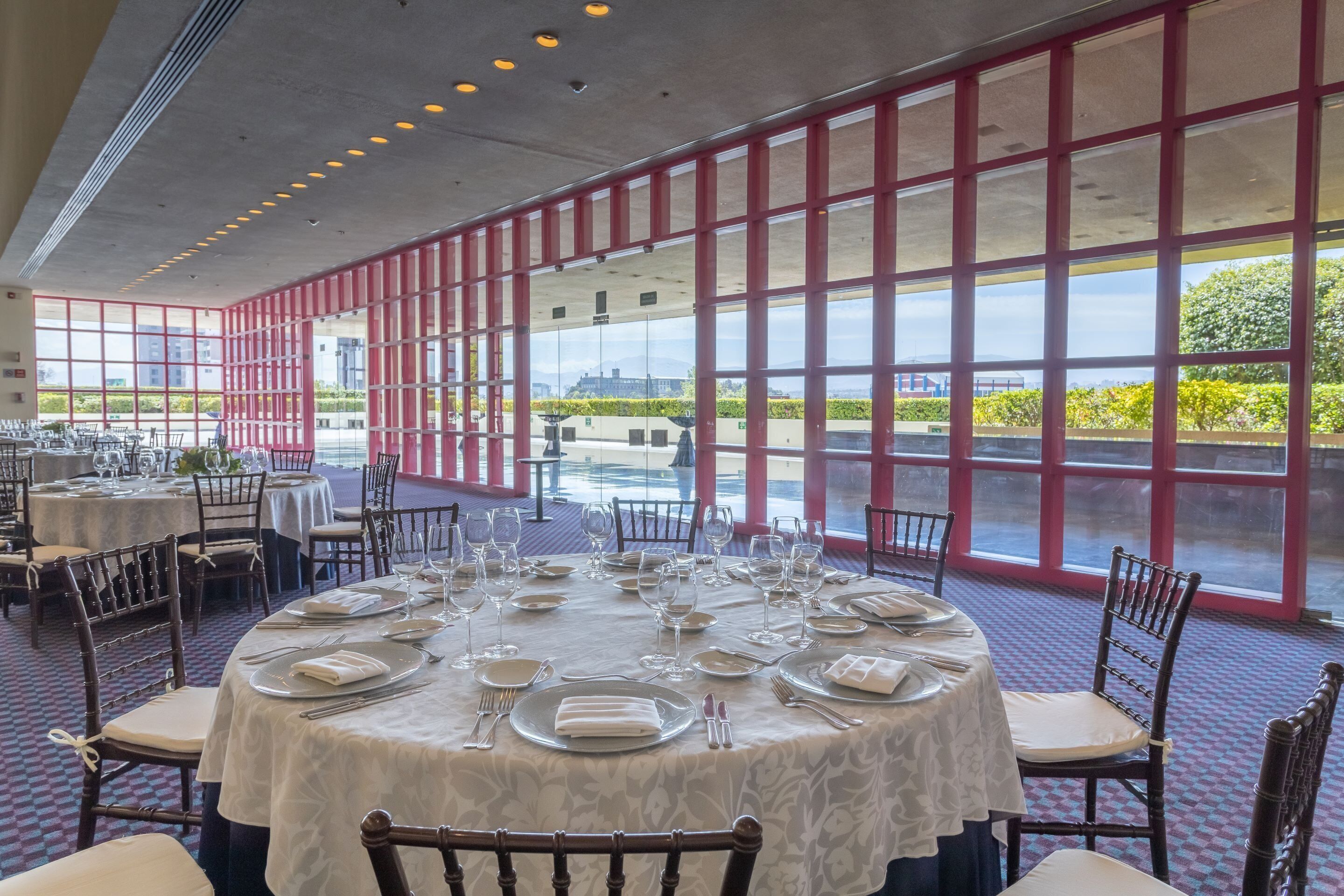 A dining area with round tables set for a meal, large windows with a view of greenery and buildings, and a patterned carpet.