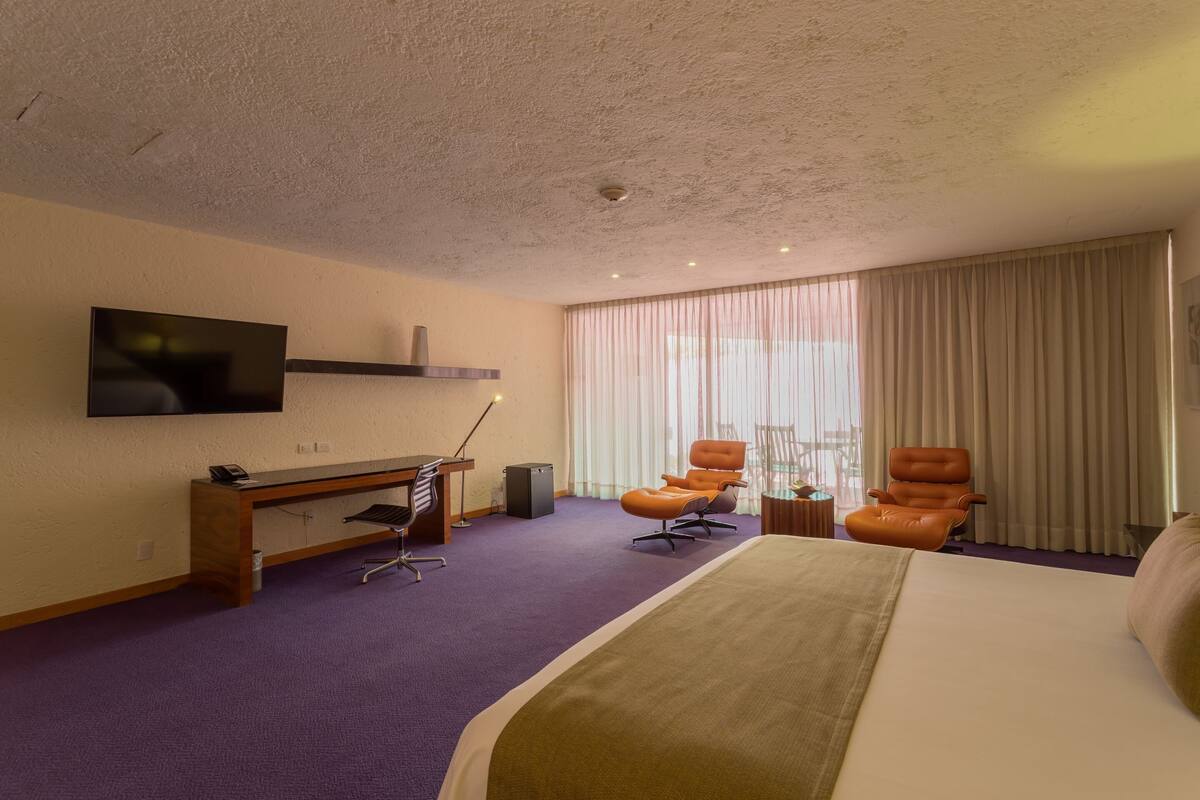 A hotel room with a bed, two armchairs, a desk with a computer, a TV mounted on the wall, and a large window with curtains.