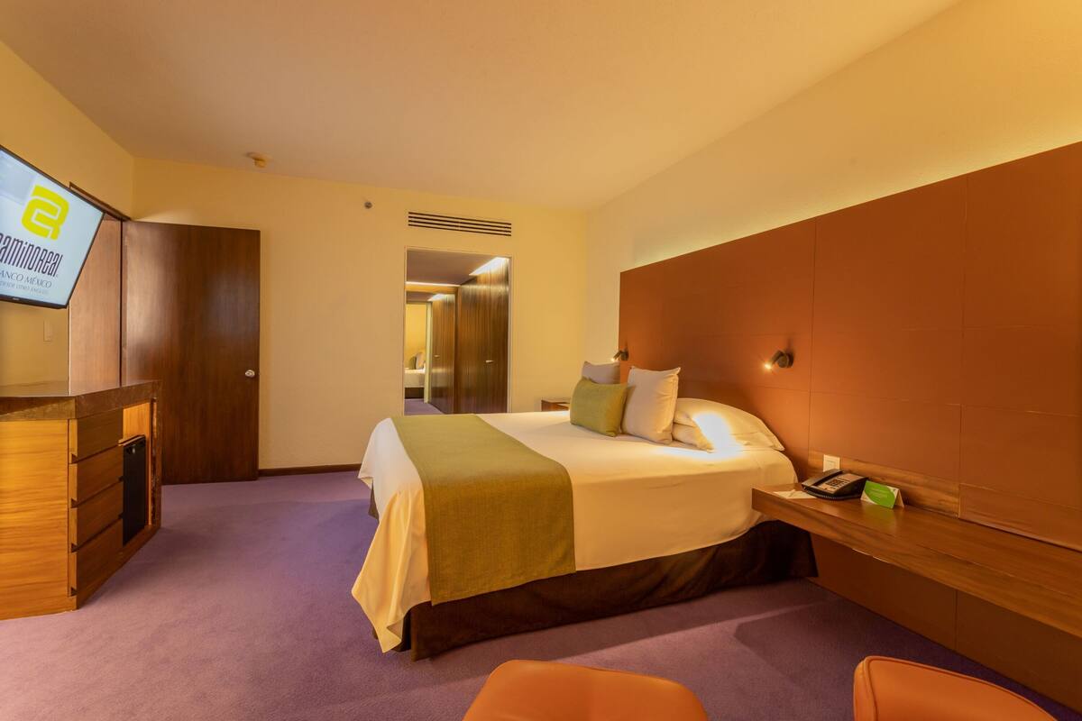 A hotel room with a bed, a desk, a chair, a TV, and a bathroom visible through an open door.