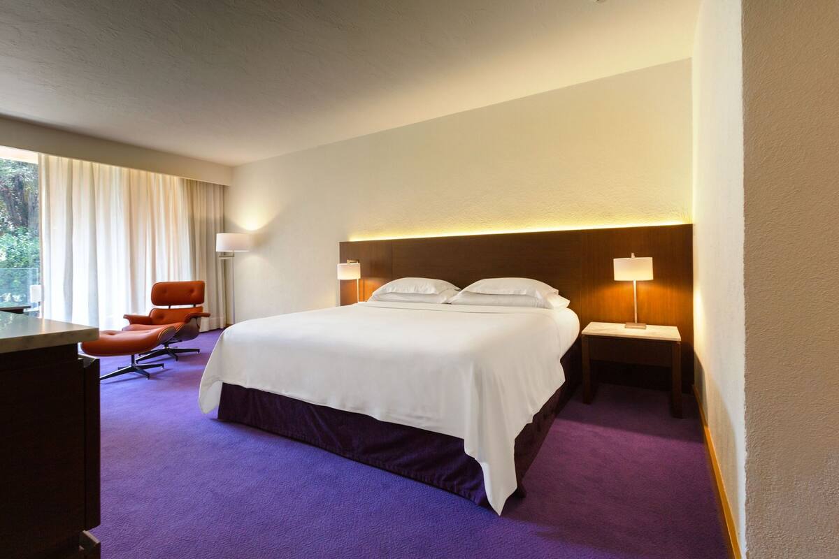 A hotel room with a large bed, two bedside tables with lamps, a desk, an orange chair, and a window with curtains.
