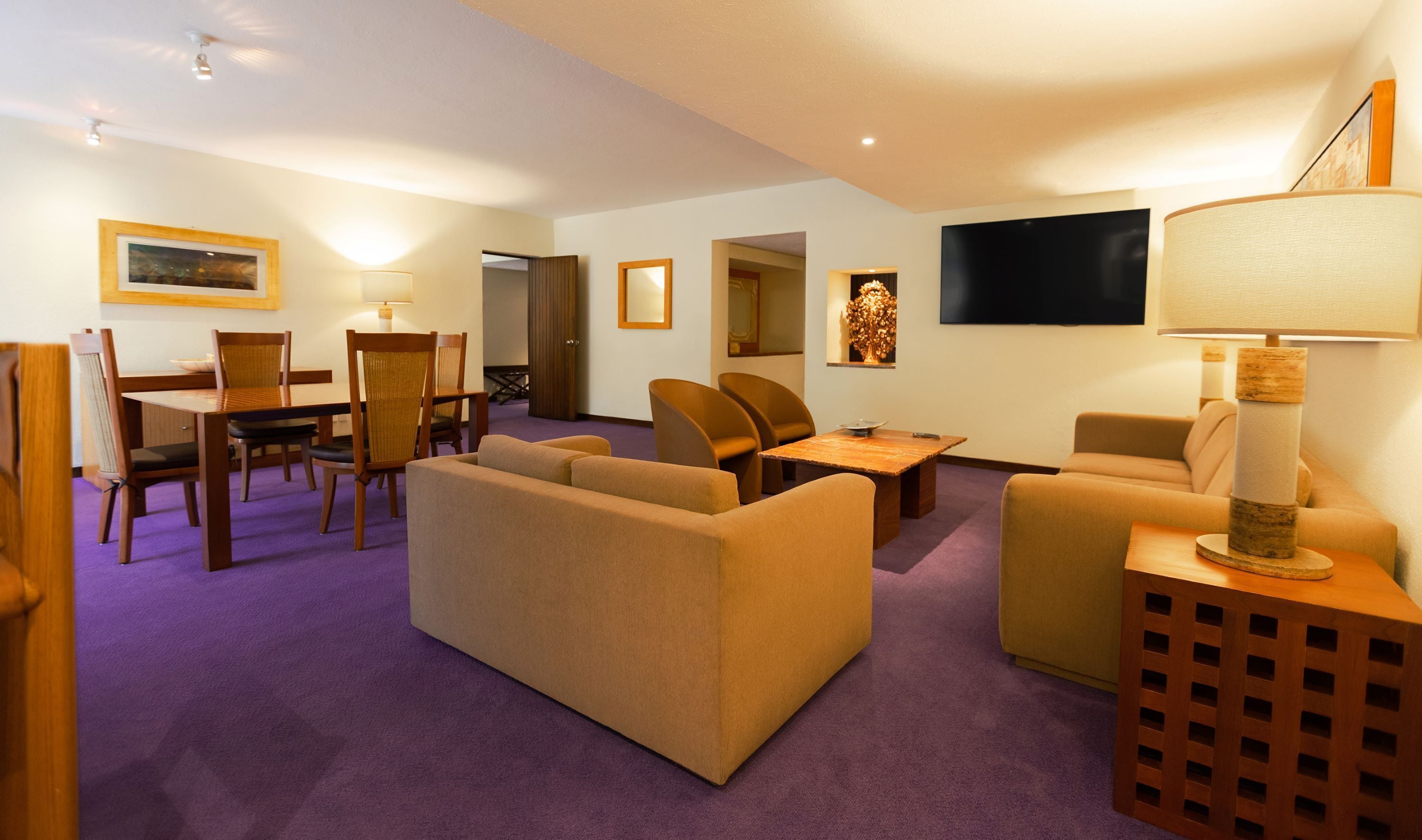 A hotel room with a purple carpet, beige sofas, a wooden dining table, and a TV mounted on the wall.