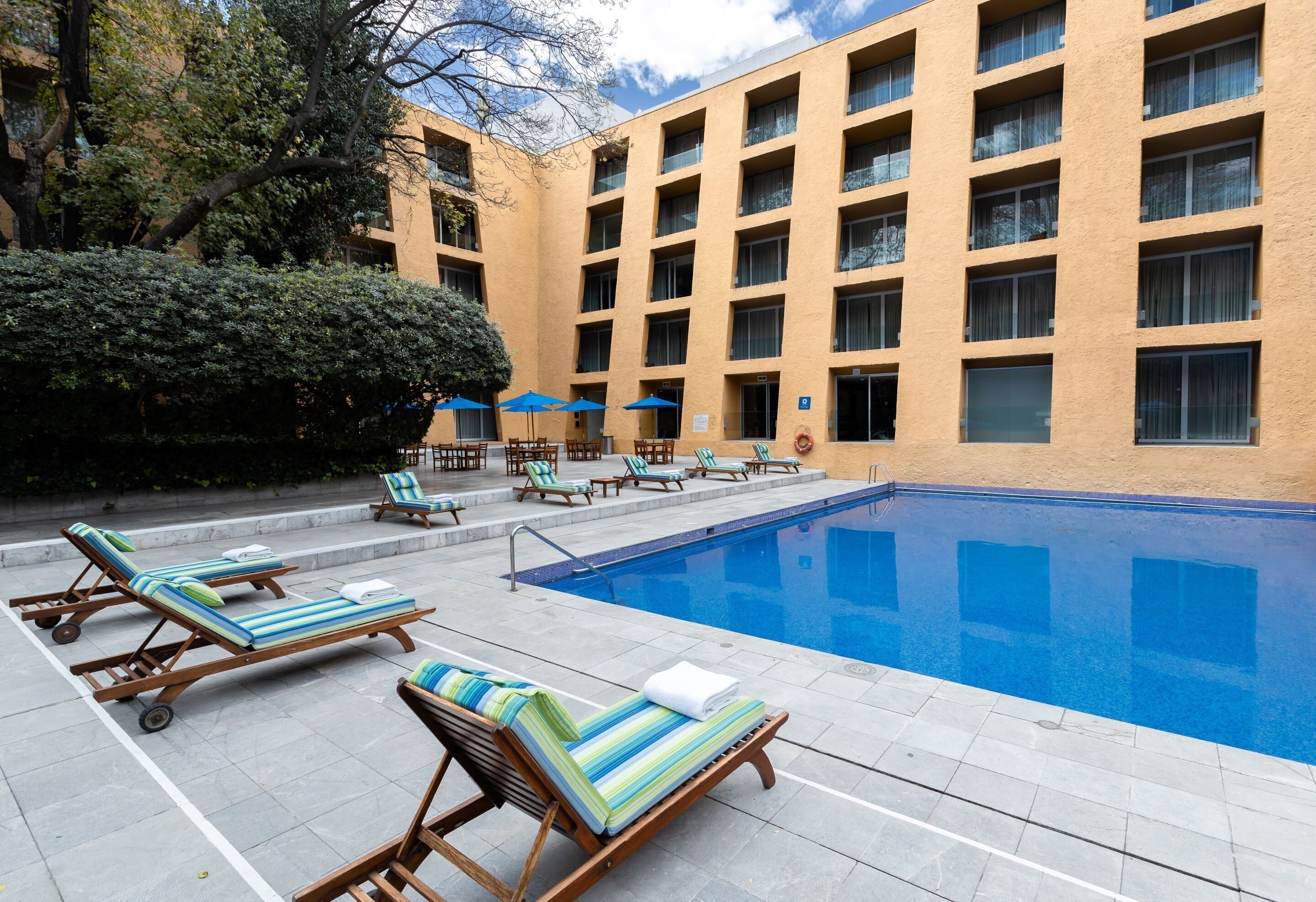 Un área de piscina con sillas de descanso y sombrillas, un edificio con múltiples ventanas y una piscina azul cristalina.