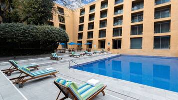 Un área de piscina con sillas de descanso y sombrillas, un edificio con múltiples ventanas y una piscina azul cristalina.