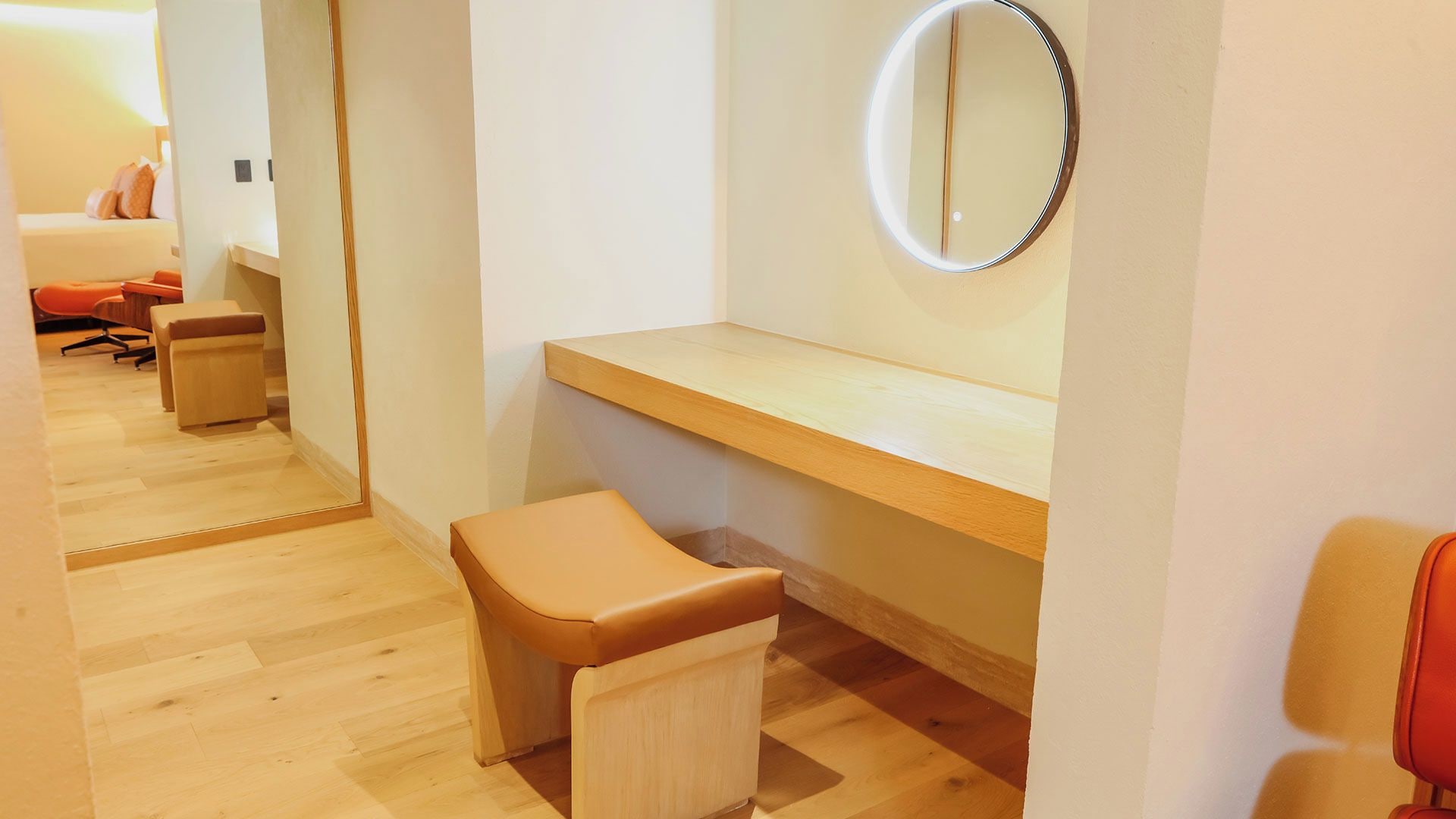 Habitación de hotel con piso de madera, un taburete pequeño, un banco de madera y un espejo en la pared.