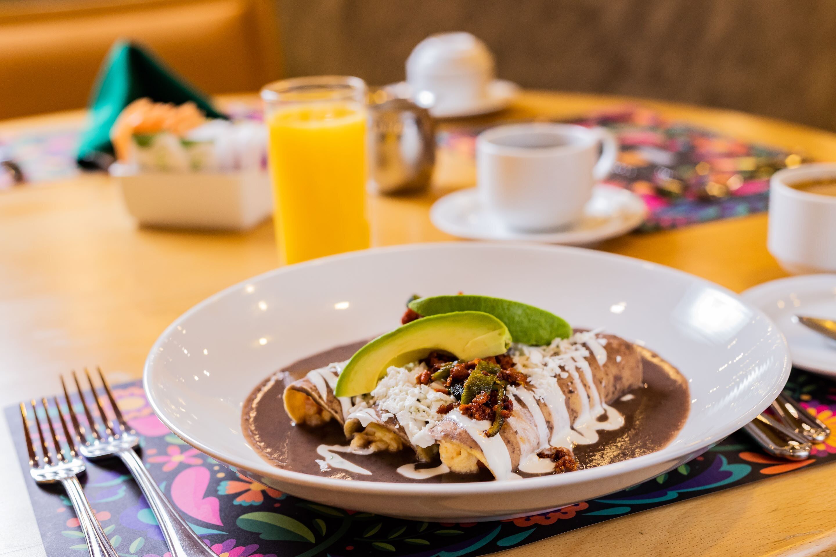 Ein Teller Enchiladas mit Avocado und Soße, ein Glas Orangensaft und eine Tasse Kaffee auf einem Tisch mit einer bunten Tischmatte.