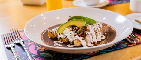 Ein Teller Enchiladas mit Avocado und Soße, ein Glas Orangensaft und eine Tasse Kaffee auf einem Tisch mit einer bunten Tischmatte.