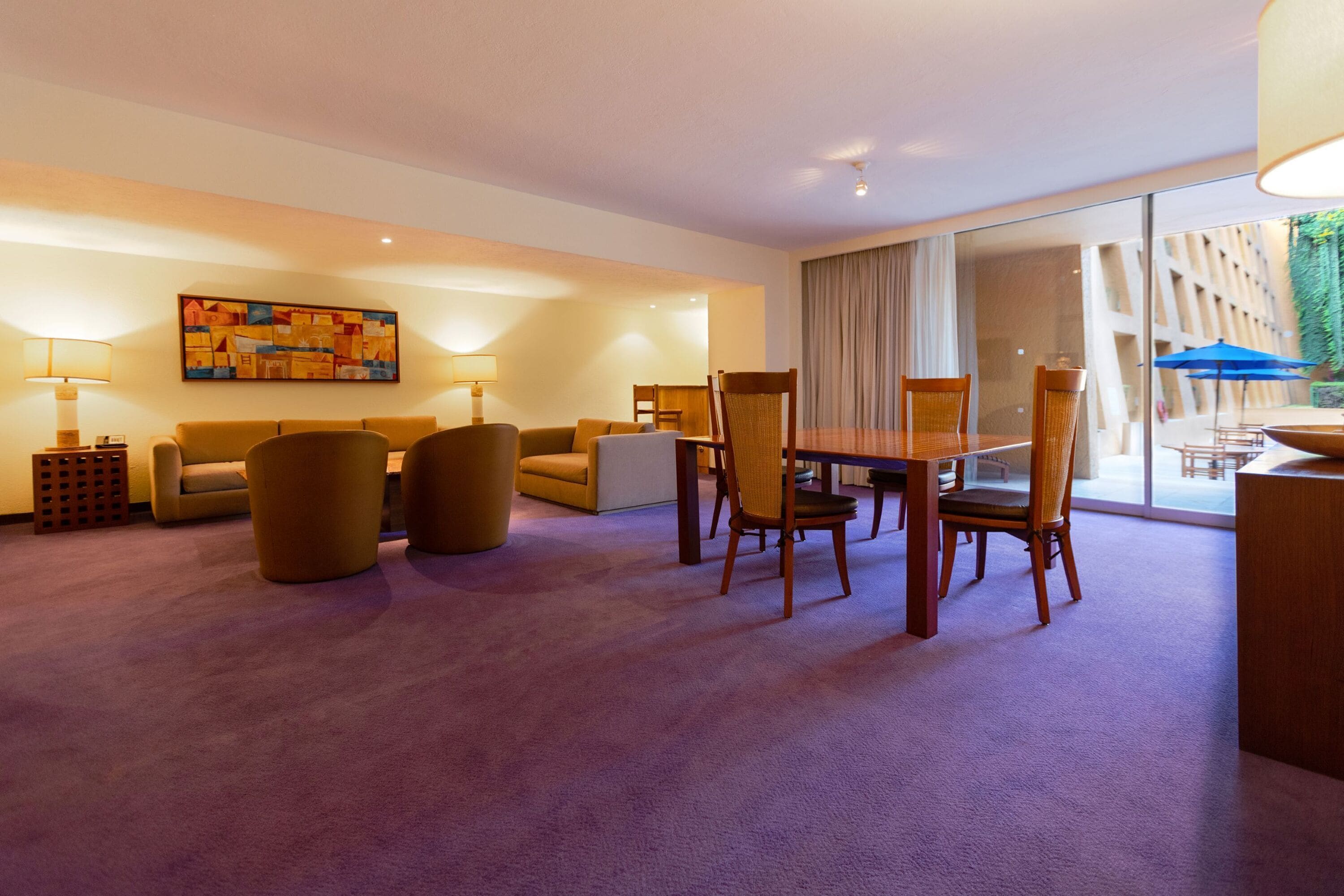 Quarto de hotel moderno com área de refeições, um conjunto de sofás e uma janela grande com cortinas.