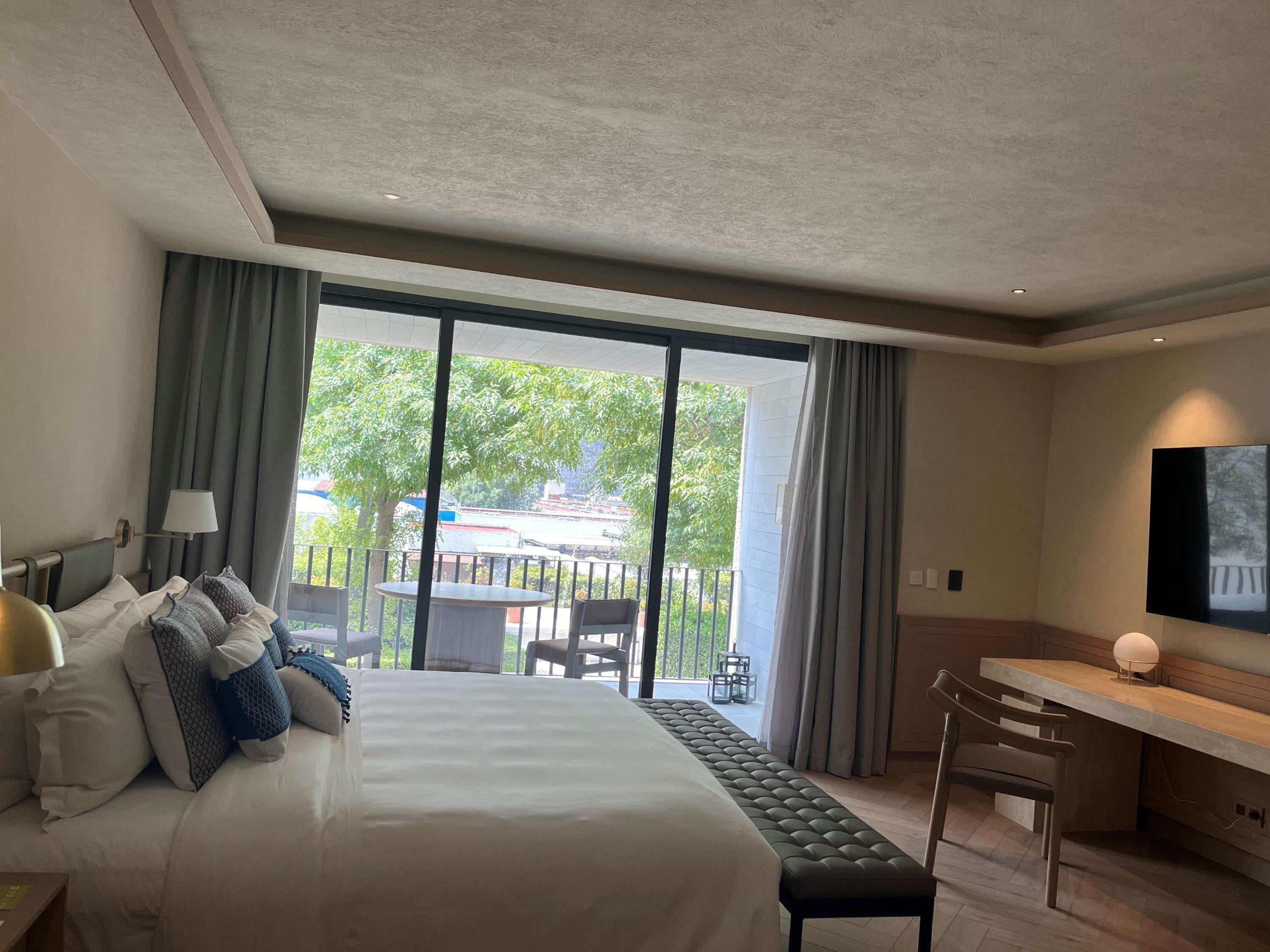 A hotel room with a bed, a desk, a chair, and a large window with a view of the outdoors.