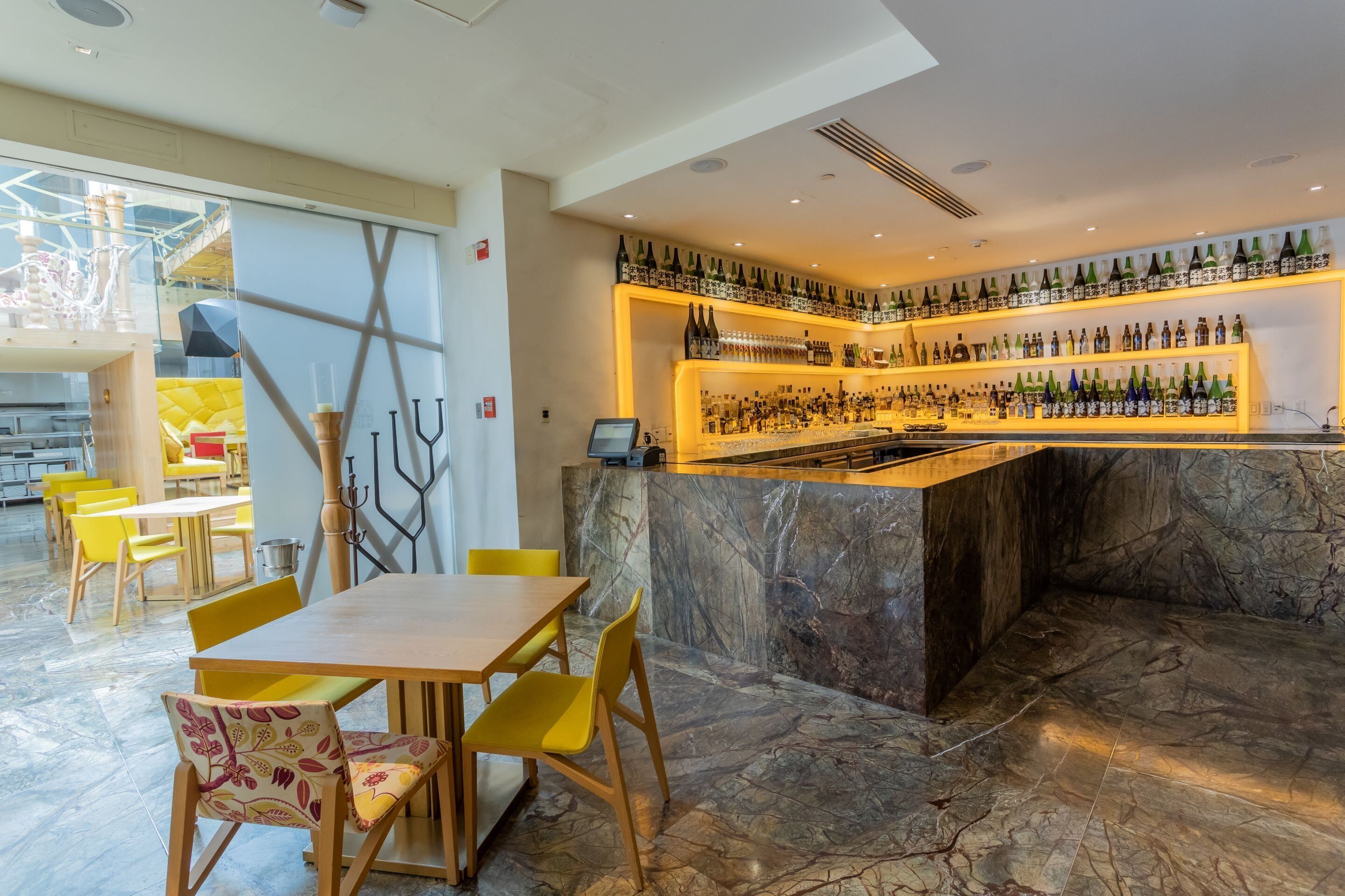 A bar area with a marble counter, yellow chairs, and a variety of bottles.