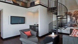 A modern living room with a flat-screen TV, grey sofas, a small kitchen area, and a spiral staircase. - Ramada Plaza by Wyndham West Hollywood Hotel & Suites (West Hollywood)