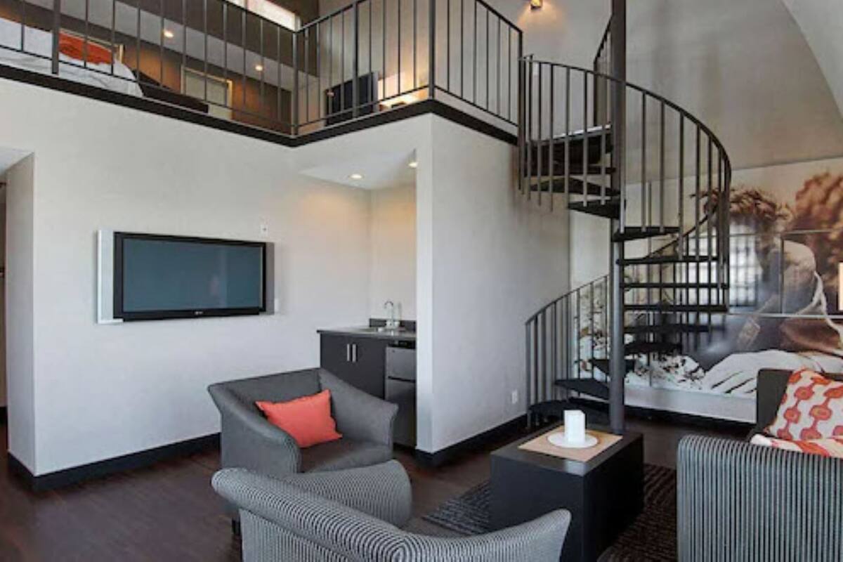 A modern living room with a flat-screen TV, grey sofas, a small kitchen area, and a spiral staircase.