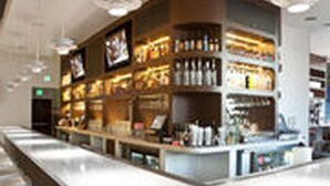 A modern bar with a curved counter, overhead lighting, and shelves stocked with bottles. - Ramada Plaza by Wyndham West Hollywood Hotel & Suites (West Hollywood)