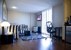 A modern living room with a dark wood floor, striped sofas, a black office chair, a flat-screen TV, and a large window with blinds.