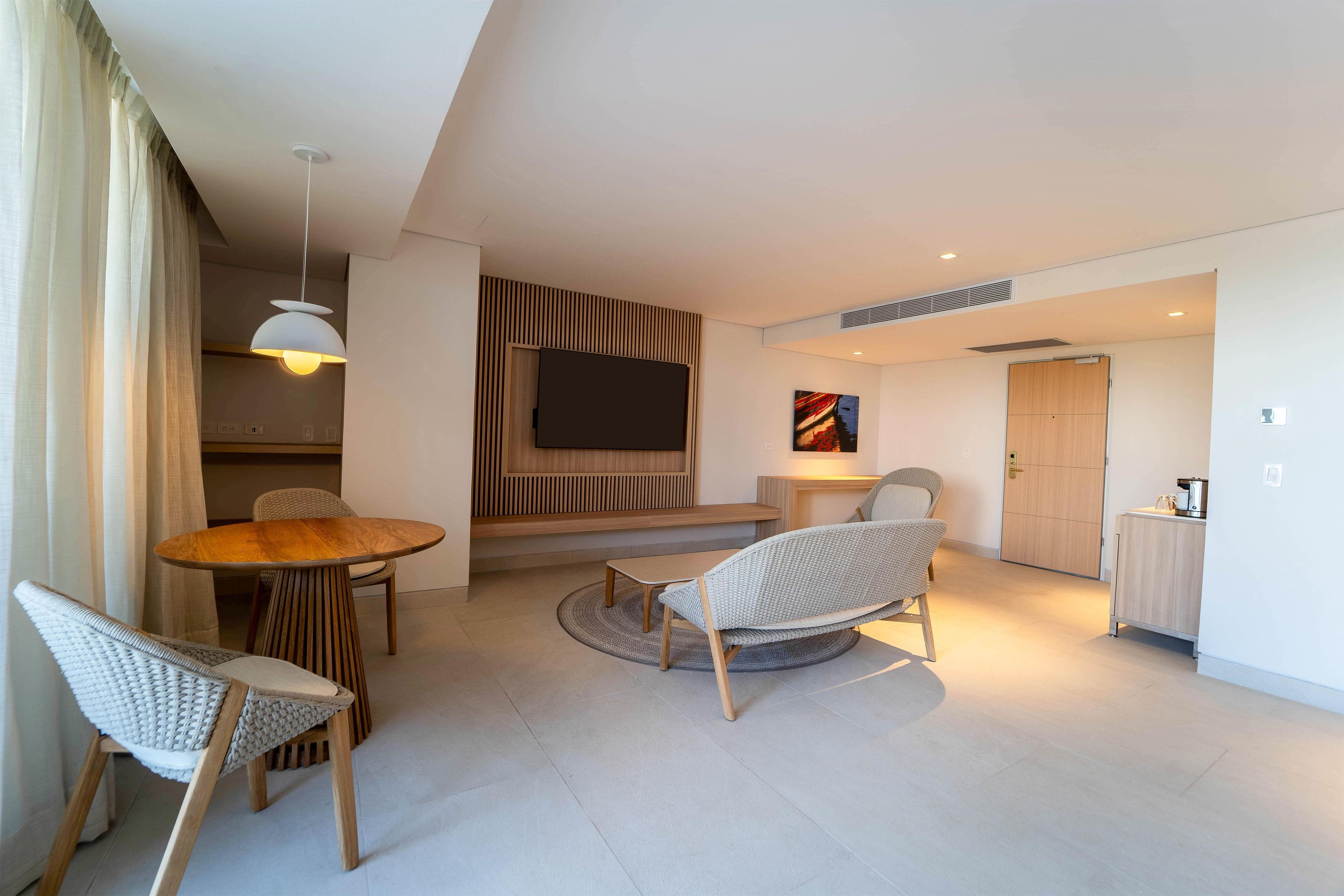 A modern living room with a round wooden table, two wicker chairs, a flat-screen TV mounted on a wooden panel, and a painting on the wall.