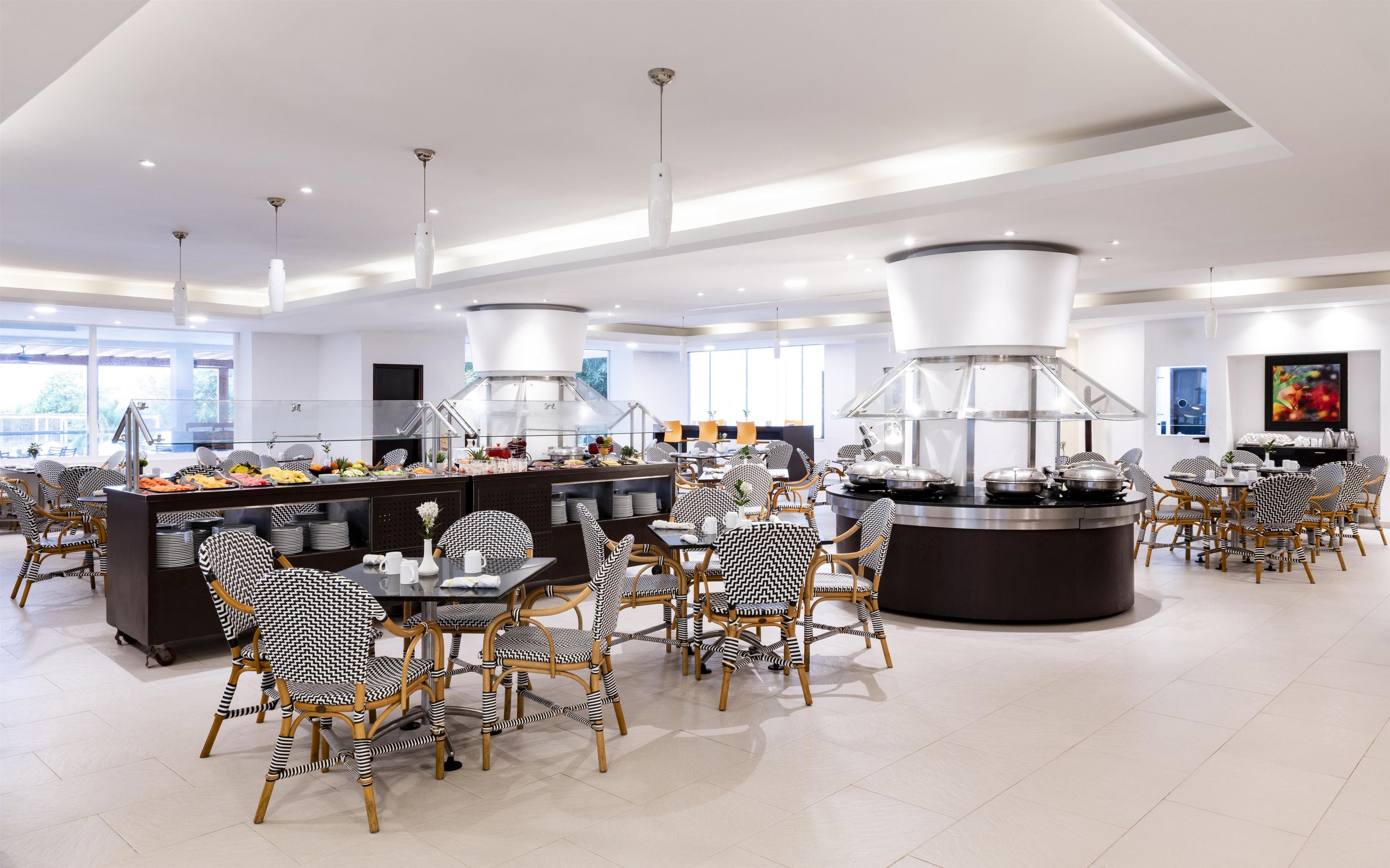 Un espace repas moderne avec des tables rondes et des chaises à motifs noir et blanc, un buffet proposant divers plats, et un grand système de ventilation de style industriel.