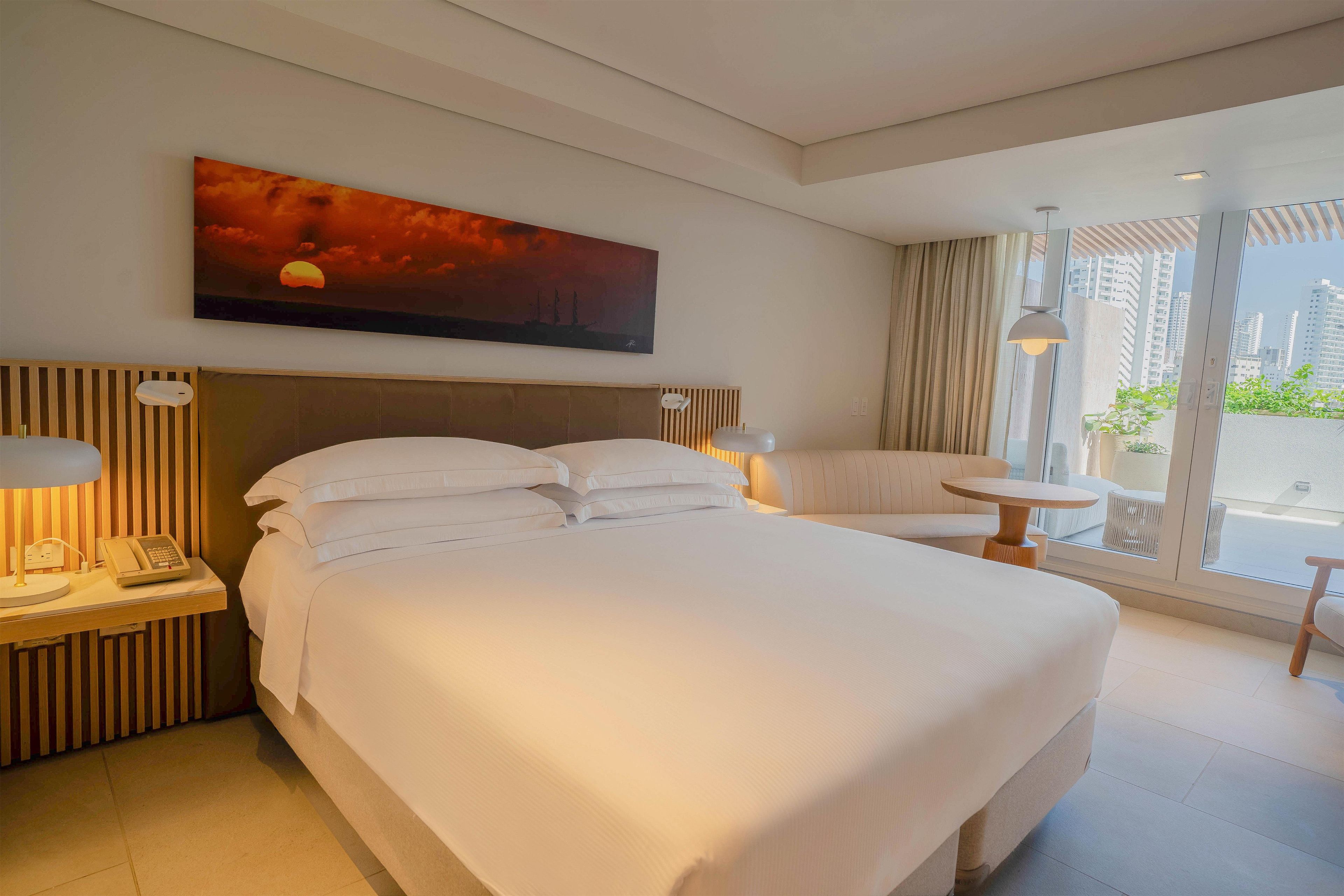 Habitación de hotel con una cama grande, mesitas de noche, un cuadro, un sofá y vistas a la ciudad.