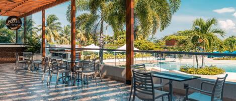 A covered outdoor seating area with tables and chairs, overlooking a pool and palm trees.