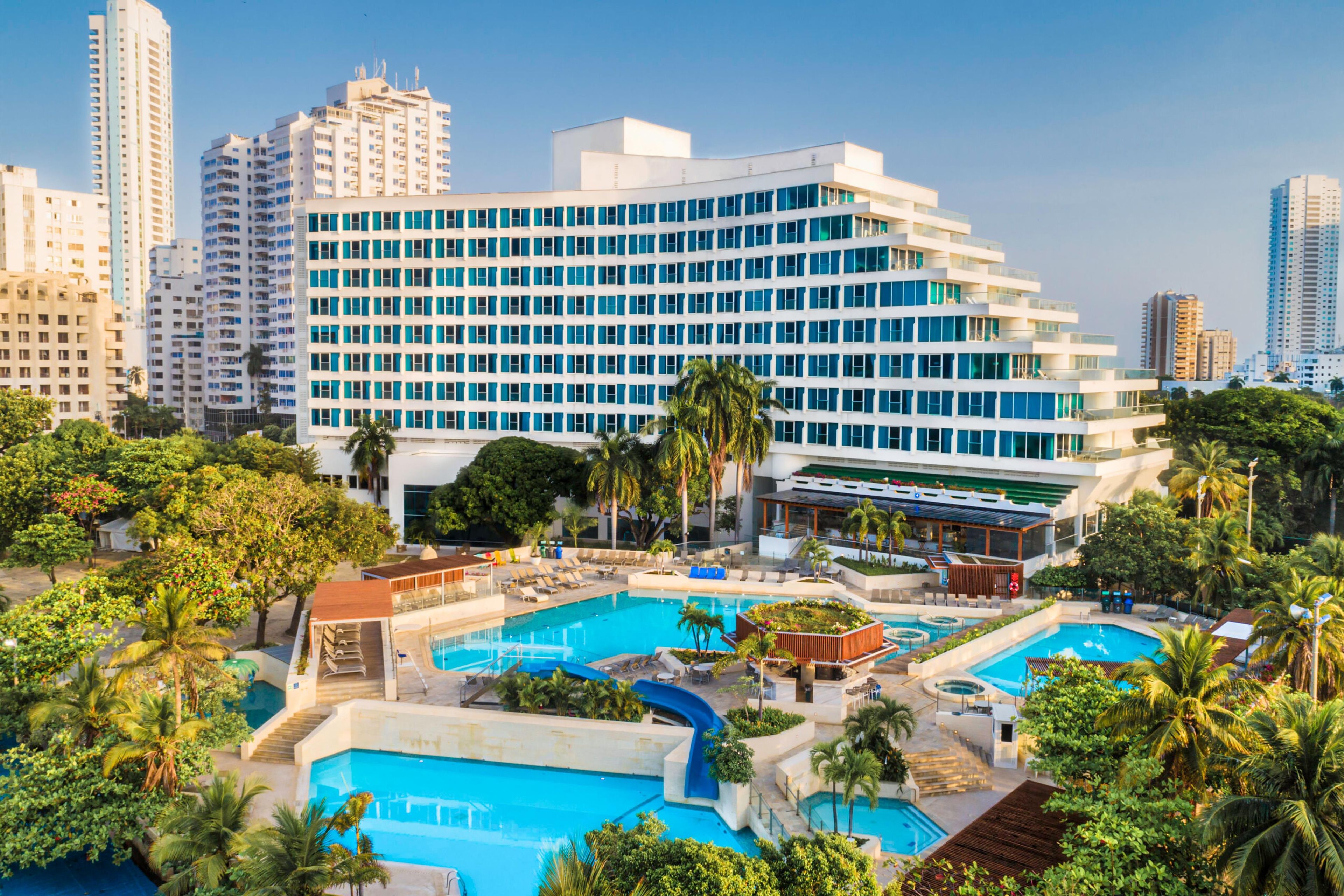 Un hotel con múltiples piscinas, un edificio amplio con muchas ventanas y vegetación circundante.