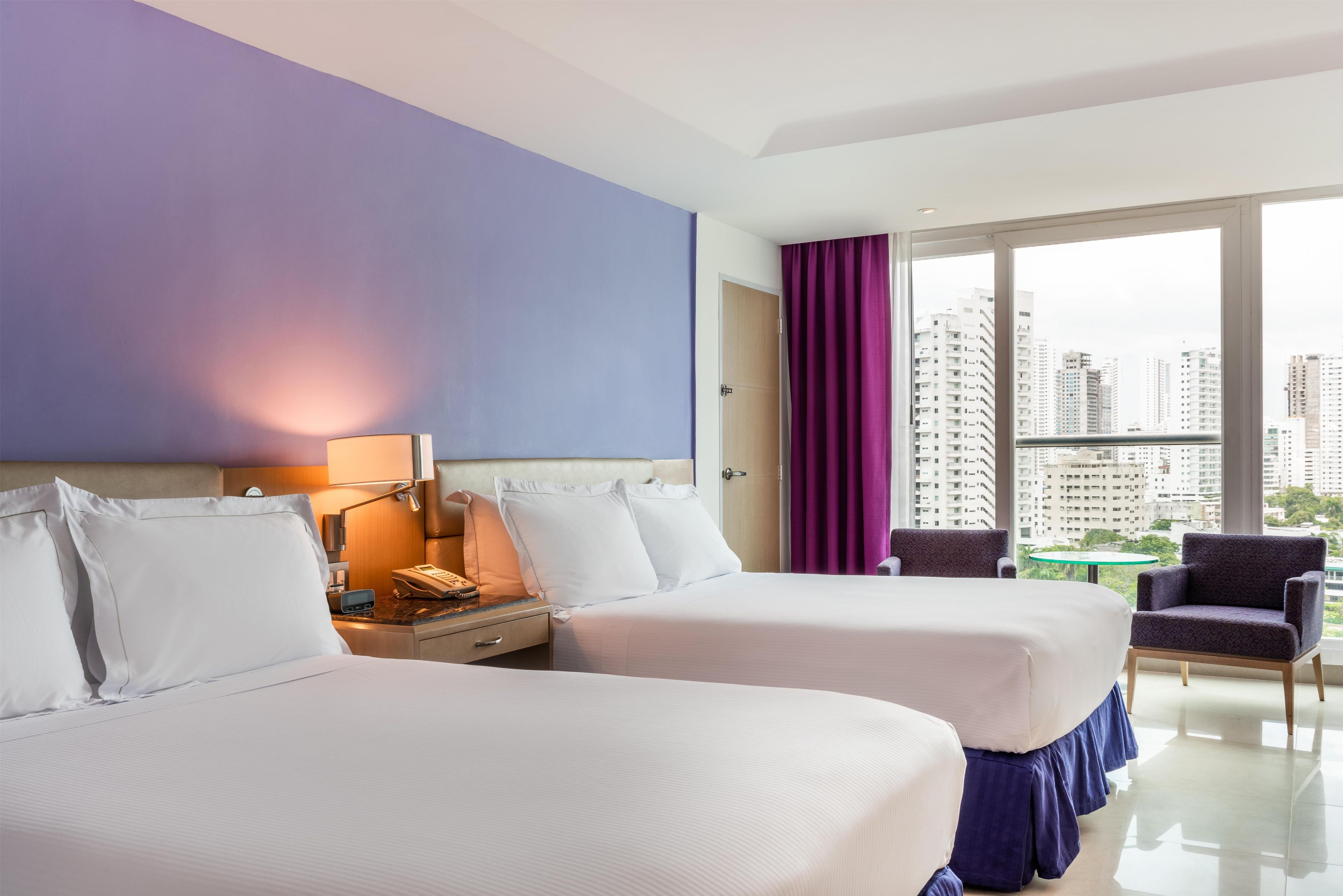 A hotel room with two beds, a nightstand, a lamp, a chair, and a view of the cityscape.