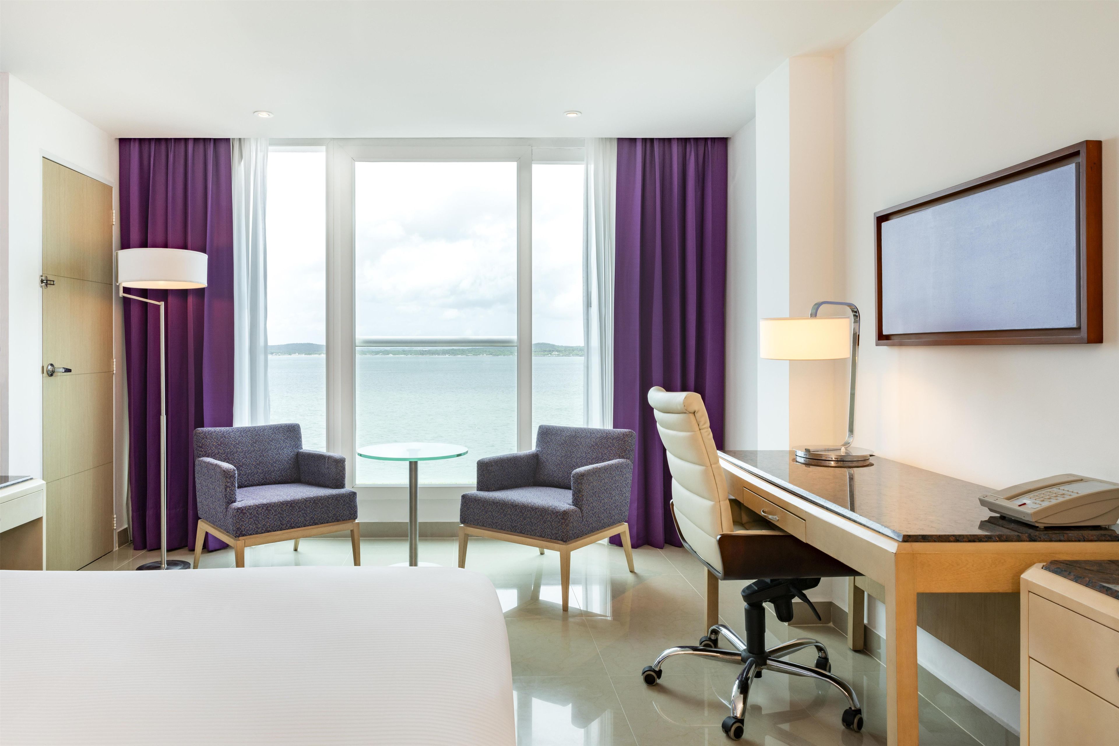 A hotel room with a bed, two armchairs, a desk with a phone, a lamp, and a view of the sea.