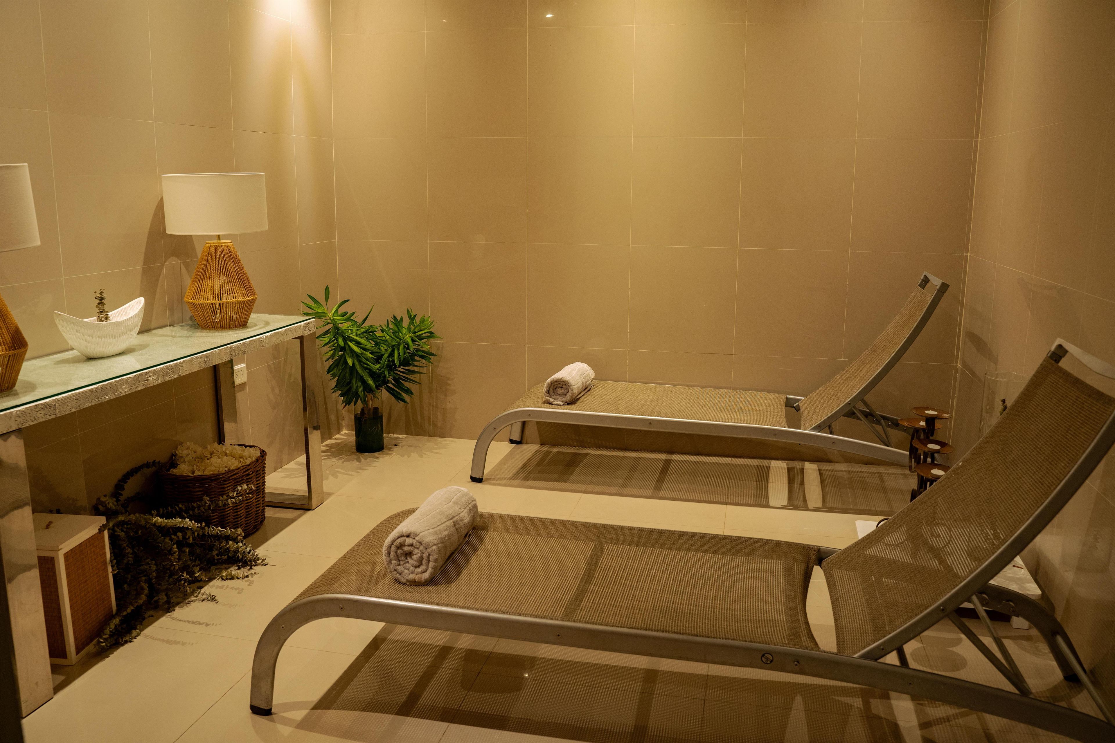 A spa room with a glass table, two massage tables, a lamp, and a potted plant.