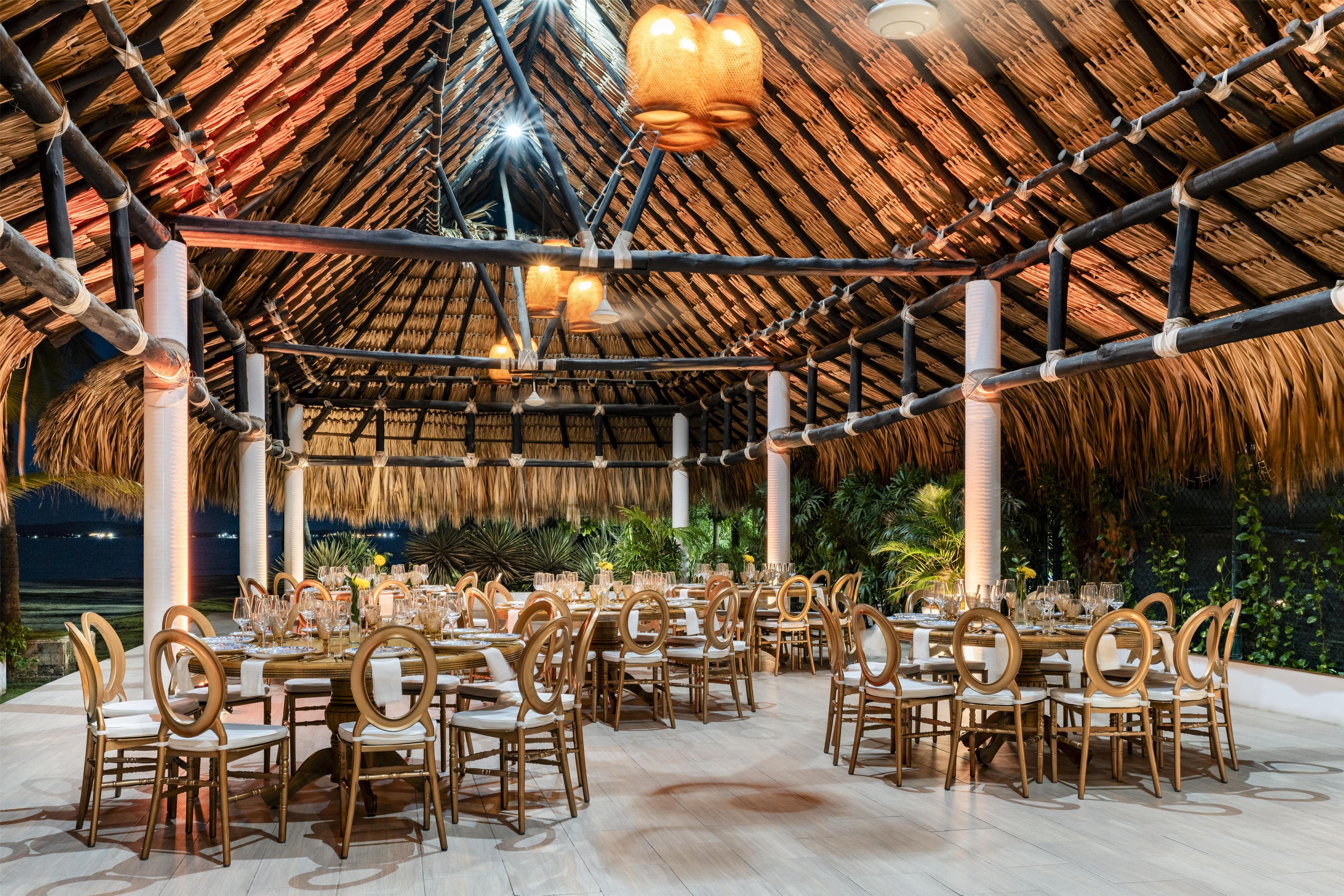 Un área de comedor al aire libre con techo de paja, sillas y mesas de madera preparadas para una comida.