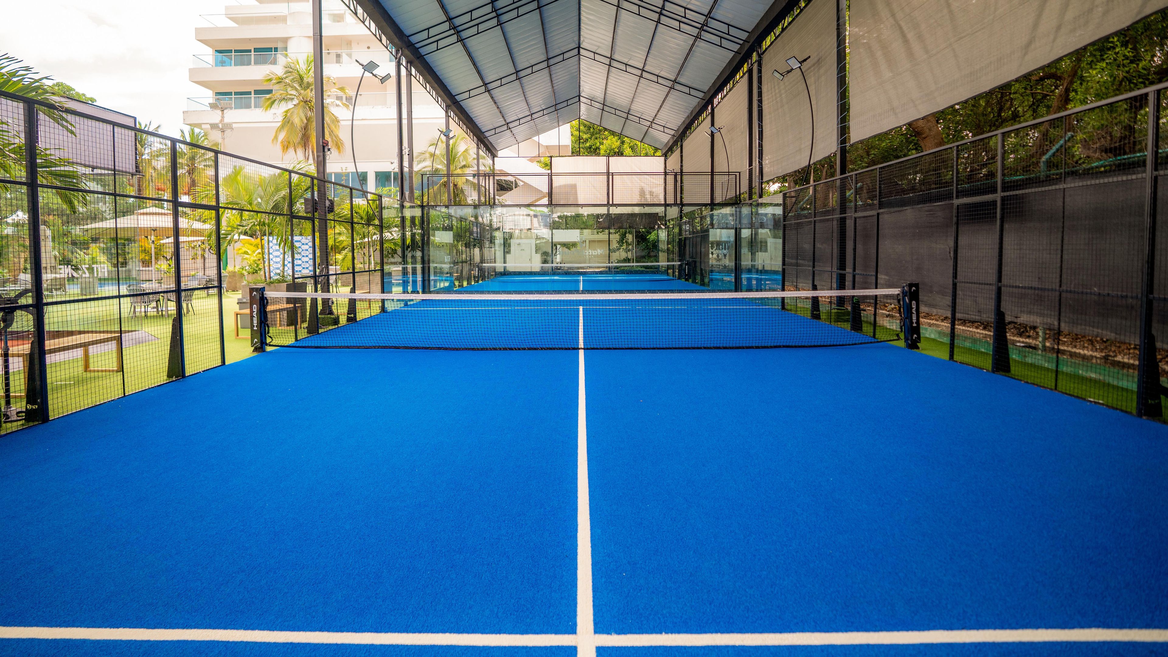 An indoor tennis court with a blue surface, surrounded by a metal fence, and a covered spectator area.