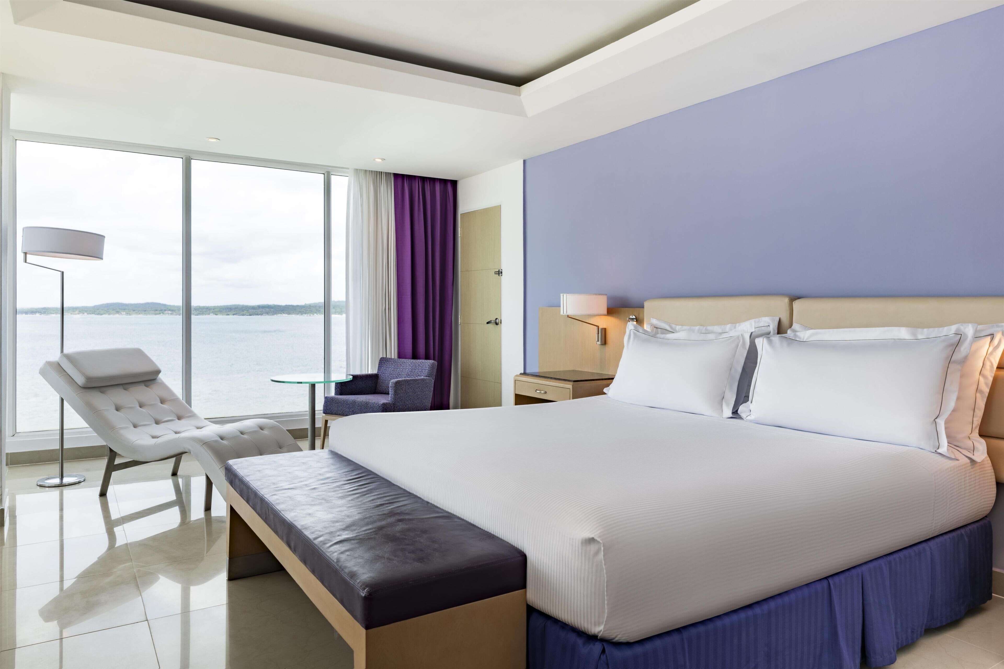 Ein modernes Hotelzimmer mit einem großen Bett, einem Lounge-Sessel, einem kleinen Tisch und Blick auf das Wasser.
