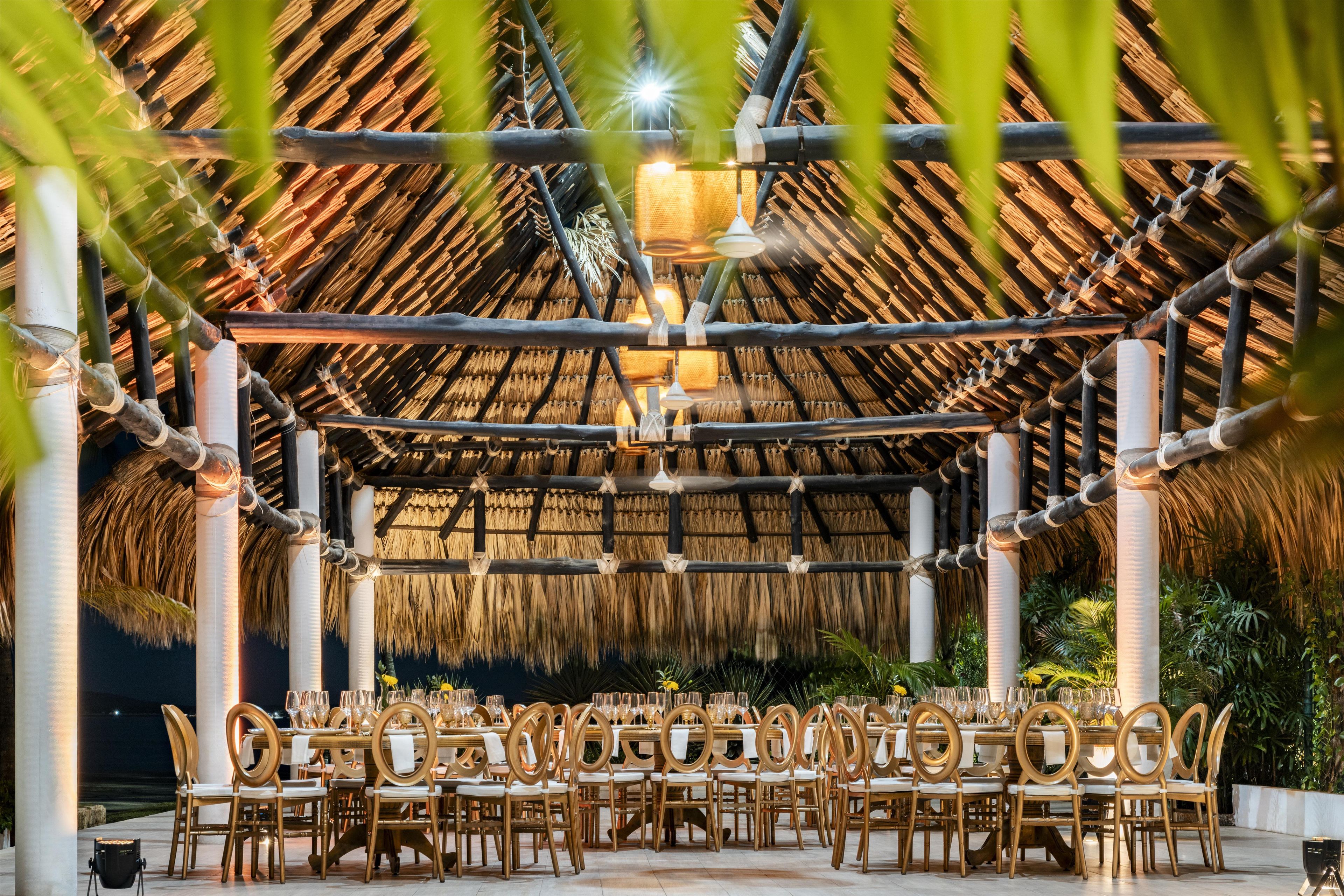 Un espacio de comedor al aire libre con techo de paja, columnas de bambú y sillas doradas.