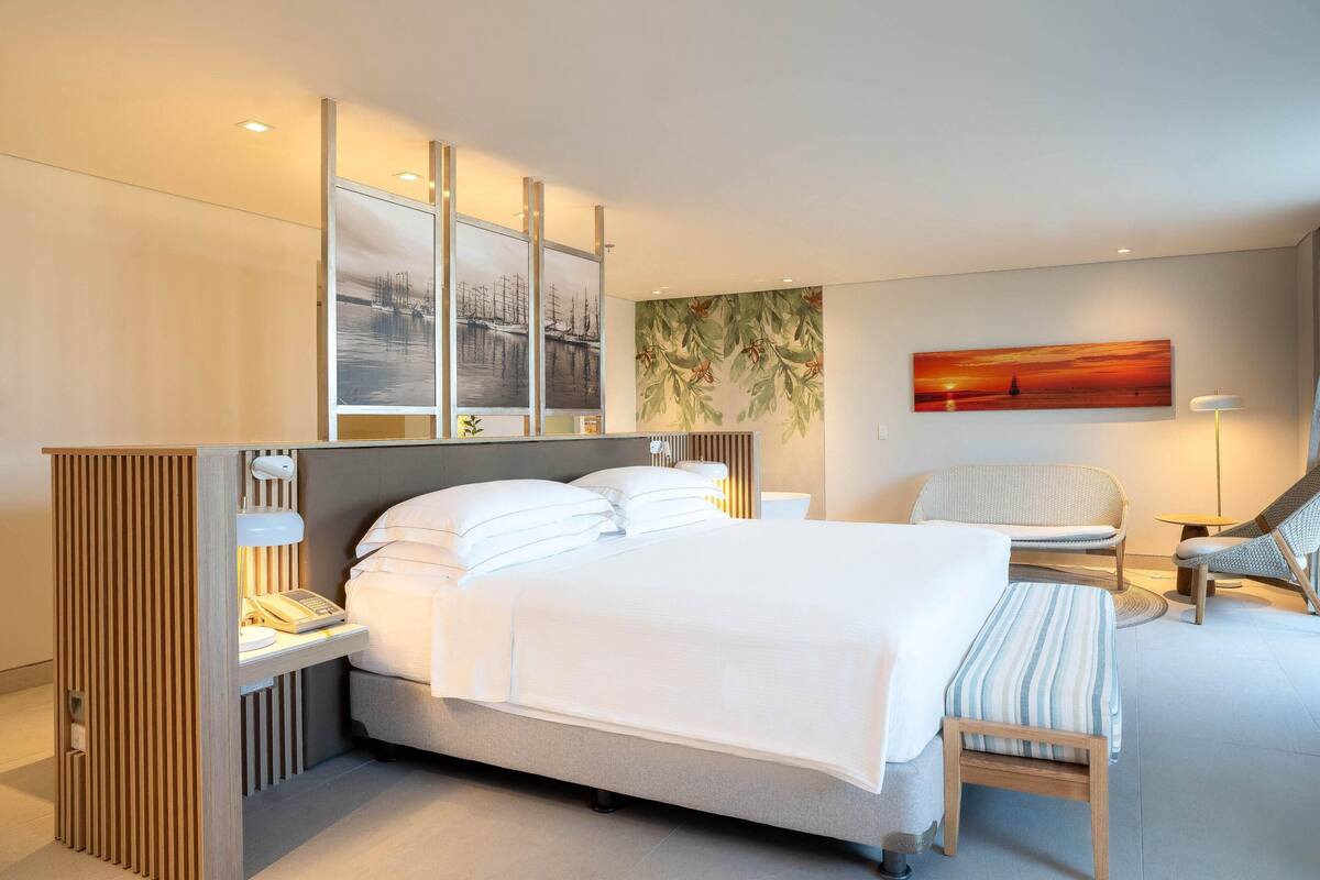 Et moderne hotelværelse med en stor seng, et natbord med lampe, en stol og kunst på væggene.
