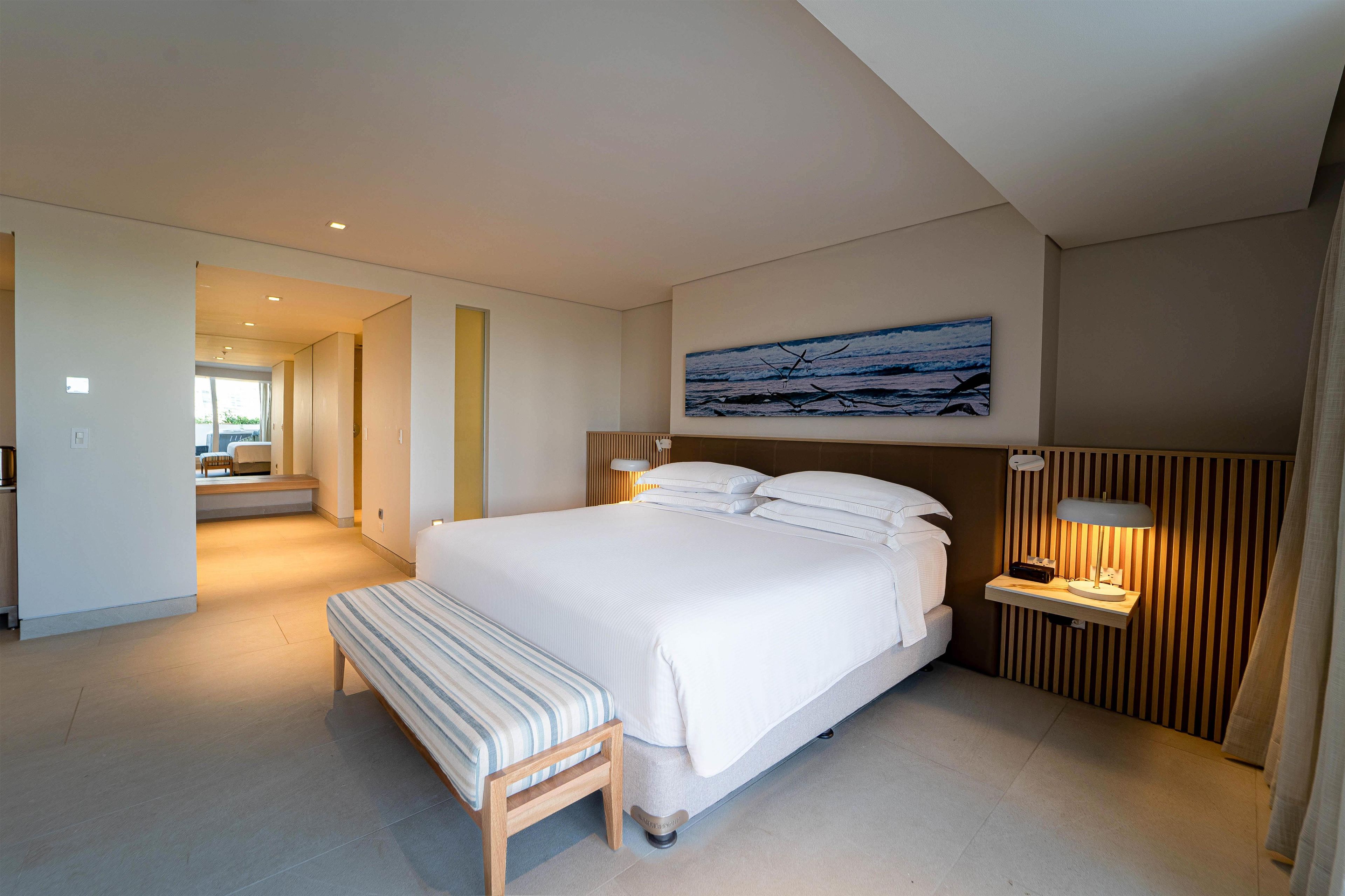 Una habitación de hotel moderna con una cama grande, una mesita de noche con lámpara, un banco y un cuadro con vista al océano.