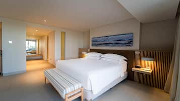 Una habitación de hotel moderna con una cama grande, una mesita de noche con lámpara, un banco y un cuadro con vista al océano.