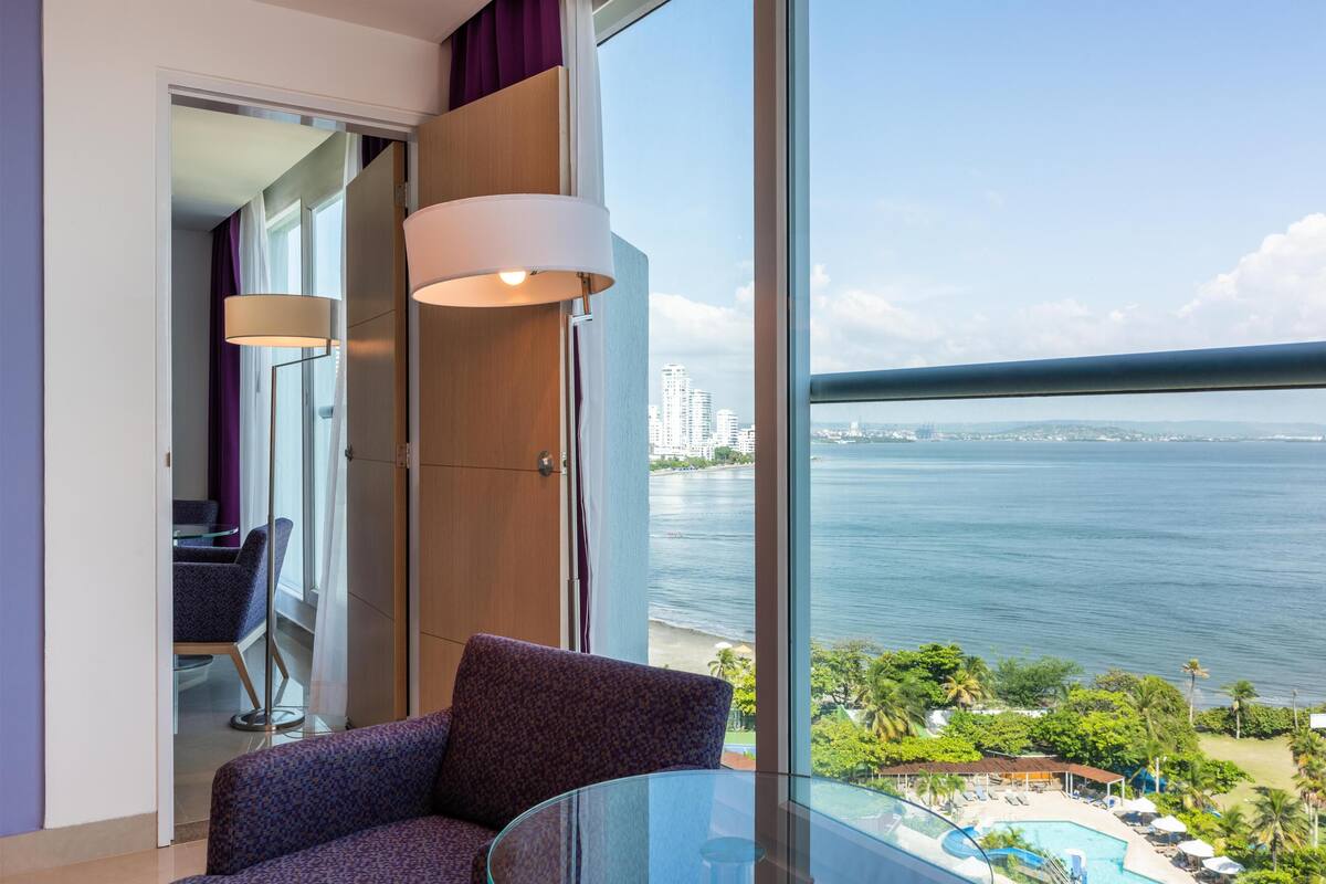 Et moderne hotelværelse med et stort vindue, et glasbord, en lilla stol og udsigt til havet og byens skyline.