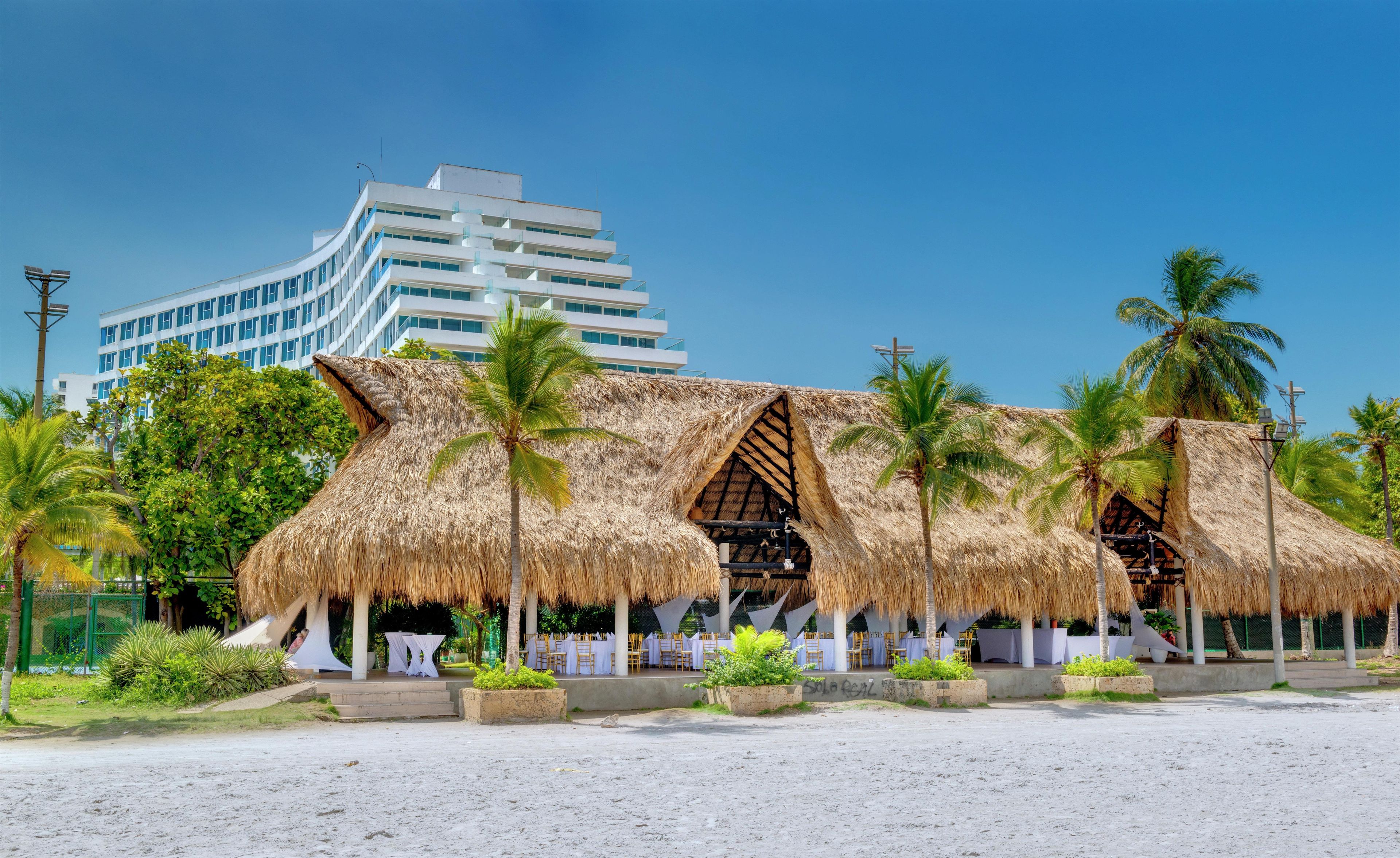 Un resort frente a la playa con estructuras de techo de paja, palmeras y un hotel moderno al fondo.