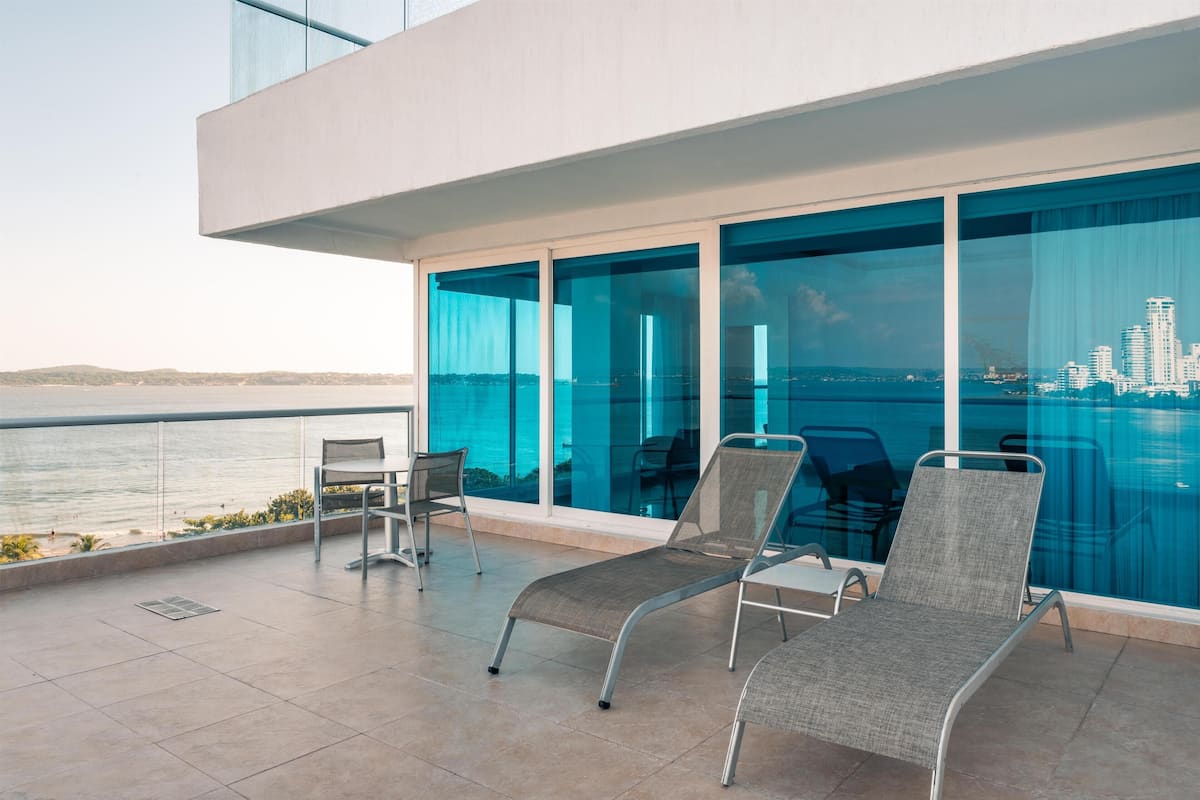 En moderne balkon med to liggestole, et bord og en stol, med udsigt over vand og byens skyline.