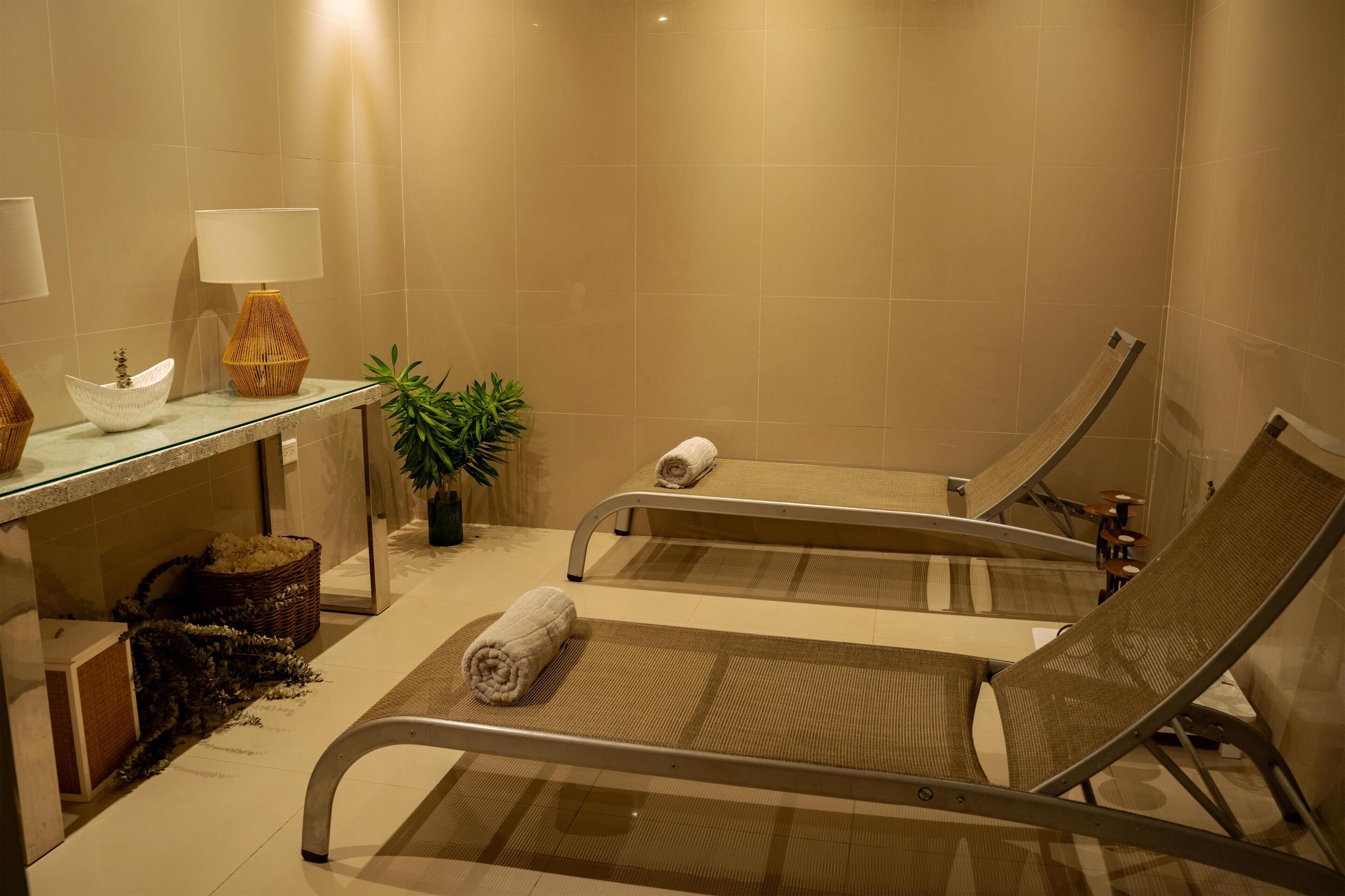 A spa room with two reclining chairs, a glass table with a lamp, a bowl, and a plant.