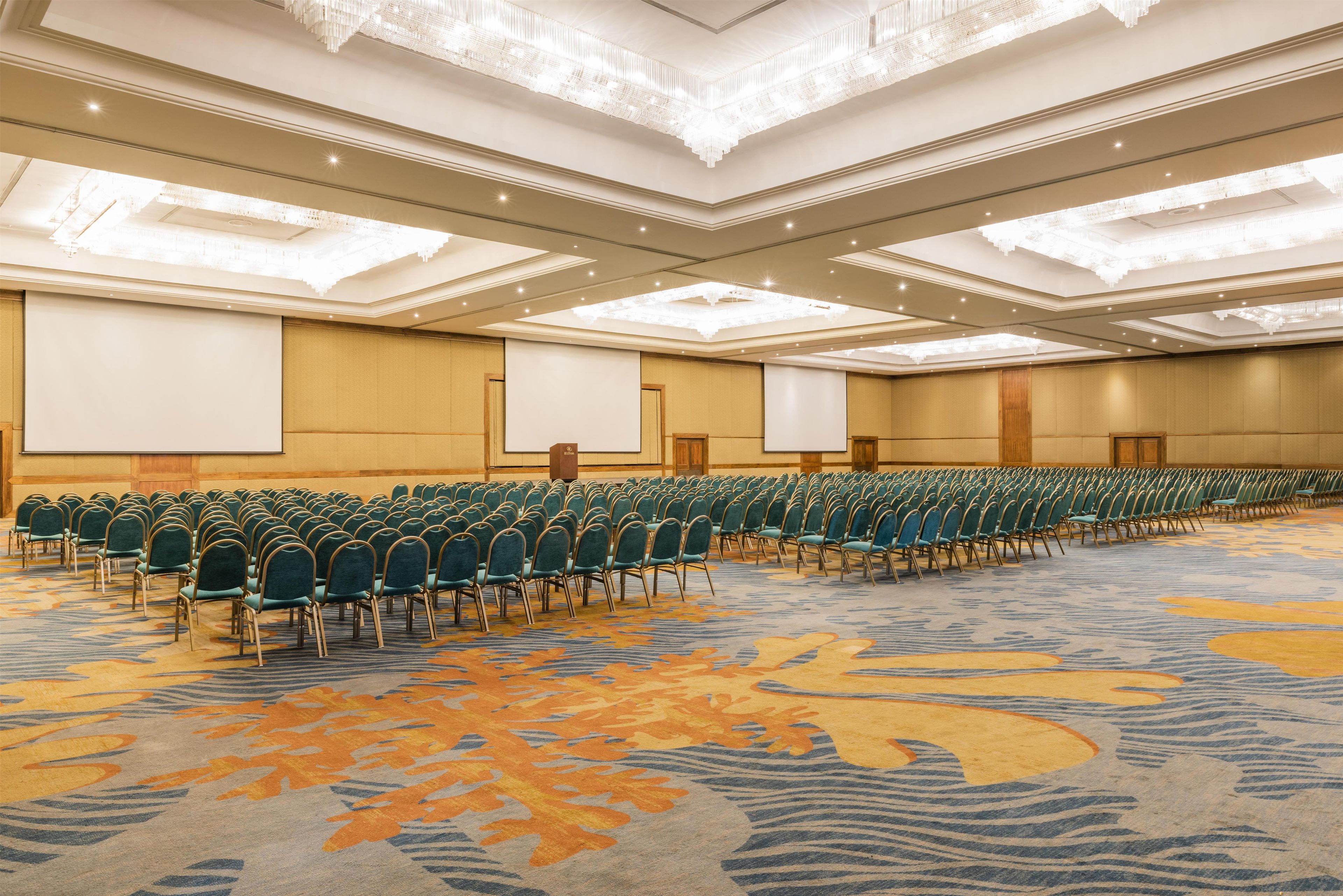 A large conference room with rows of chairs, a carpeted floor, and a high ceiling with chandeliers.