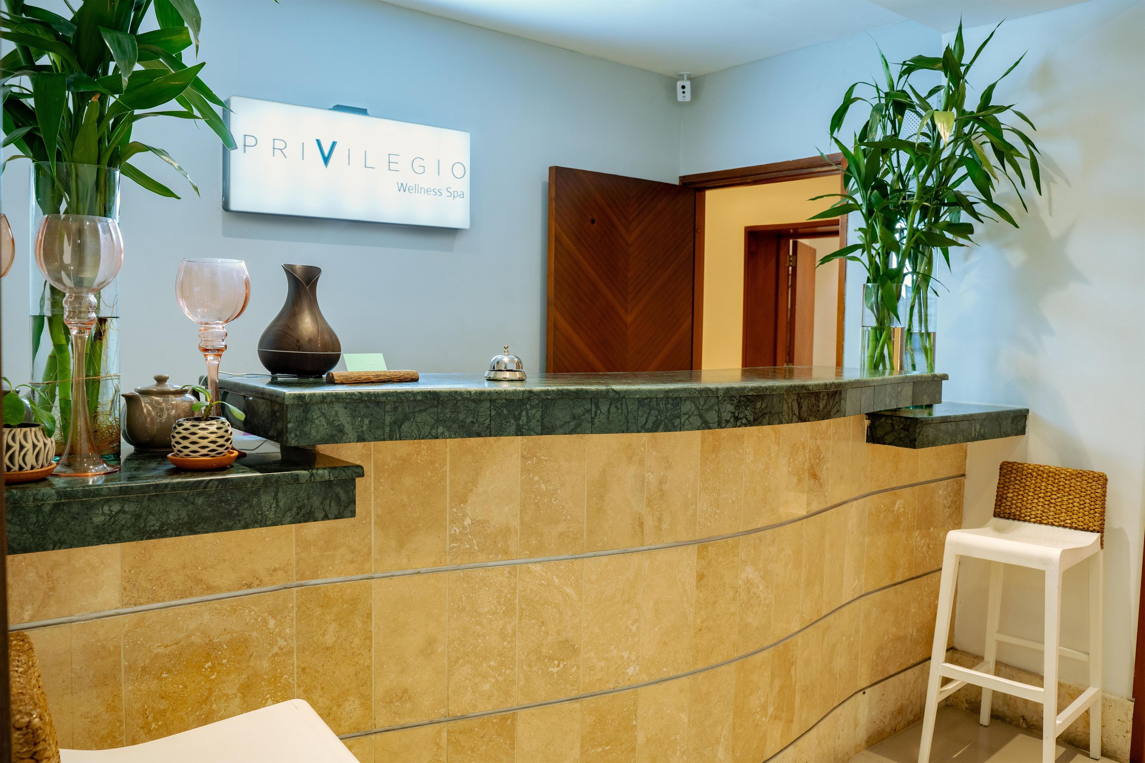 Un comptoir d’accueil avec une enseigne indiquant « PRIVILEGIO Wellness Spa », une plante en pot, un vase et une petite table avec une théière et des tasses.
