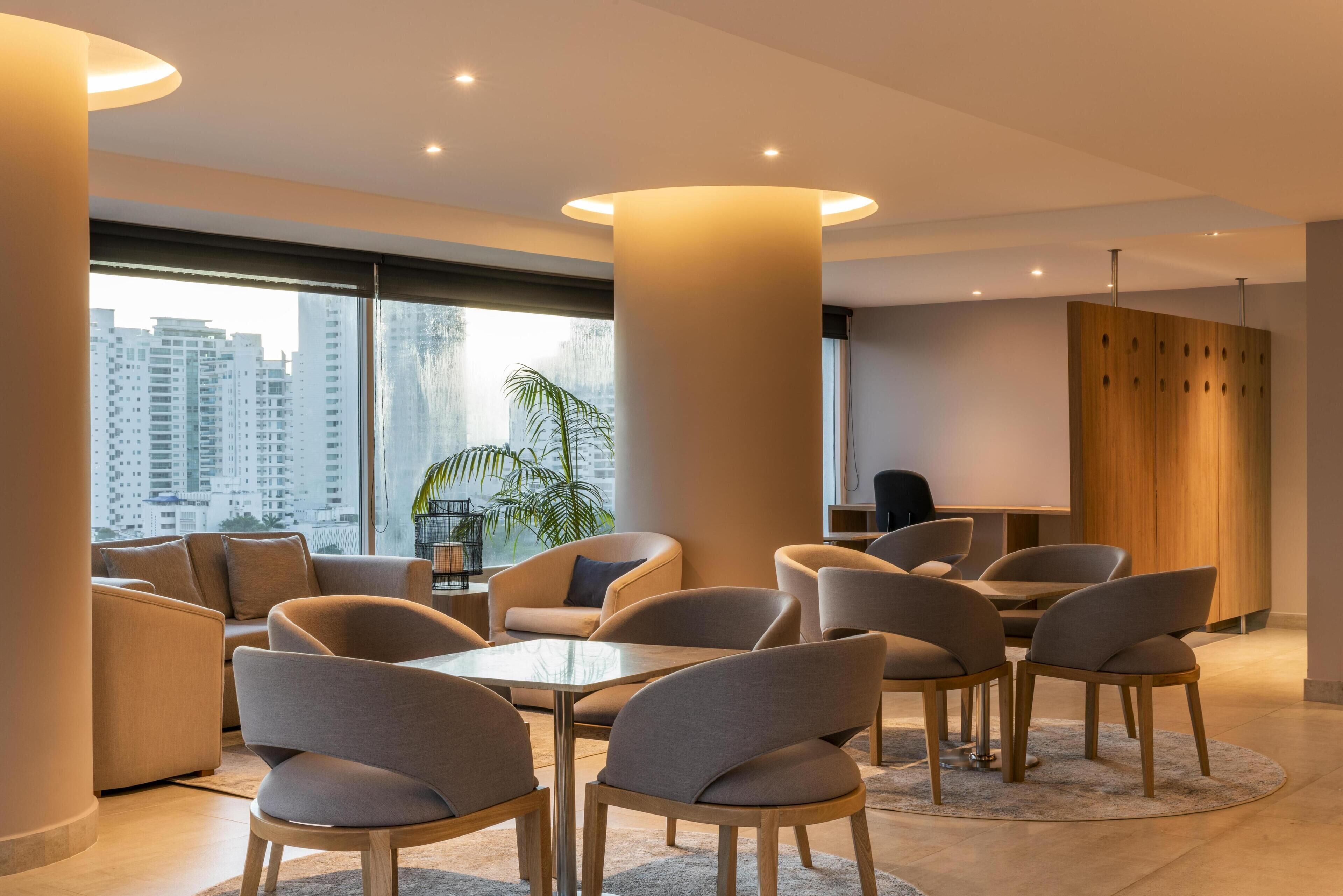 Un moderno hall de hotel con una mesa redonda, sofás y un amplio ventanal con vistas a la ciudad.