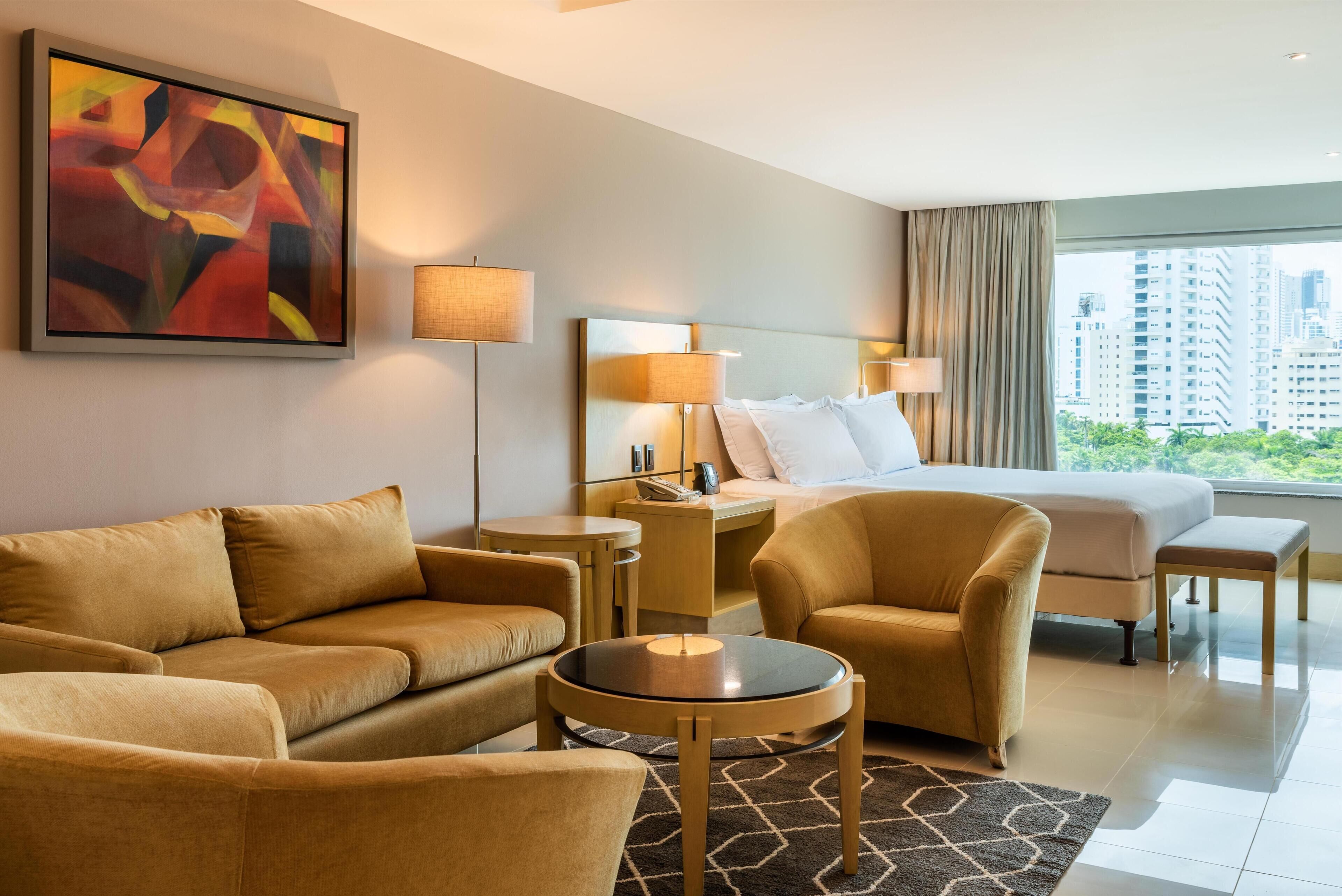 Habitación de hotel con cama, dos sofás, una mesita, una mesita de noche y un cuadro abstracto grande.