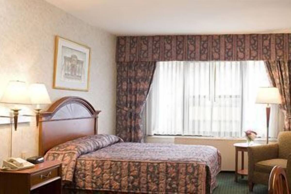 A hotel room with a bed, two bedside lamps, a chair, a small table, and a window with curtains.