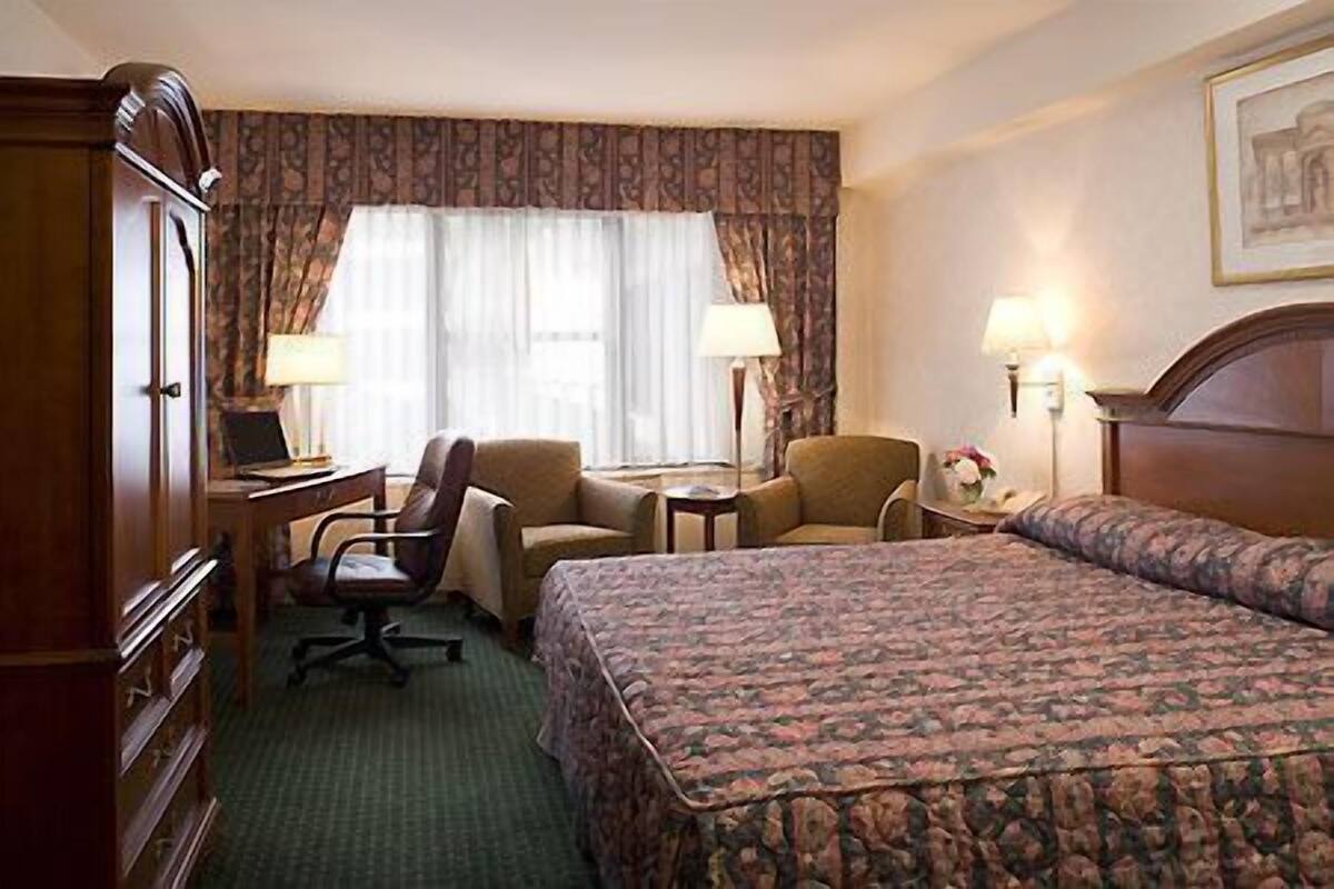 A hotel room with a bed, two armchairs, a desk with a lamp, a wardrobe, and a window with curtains.