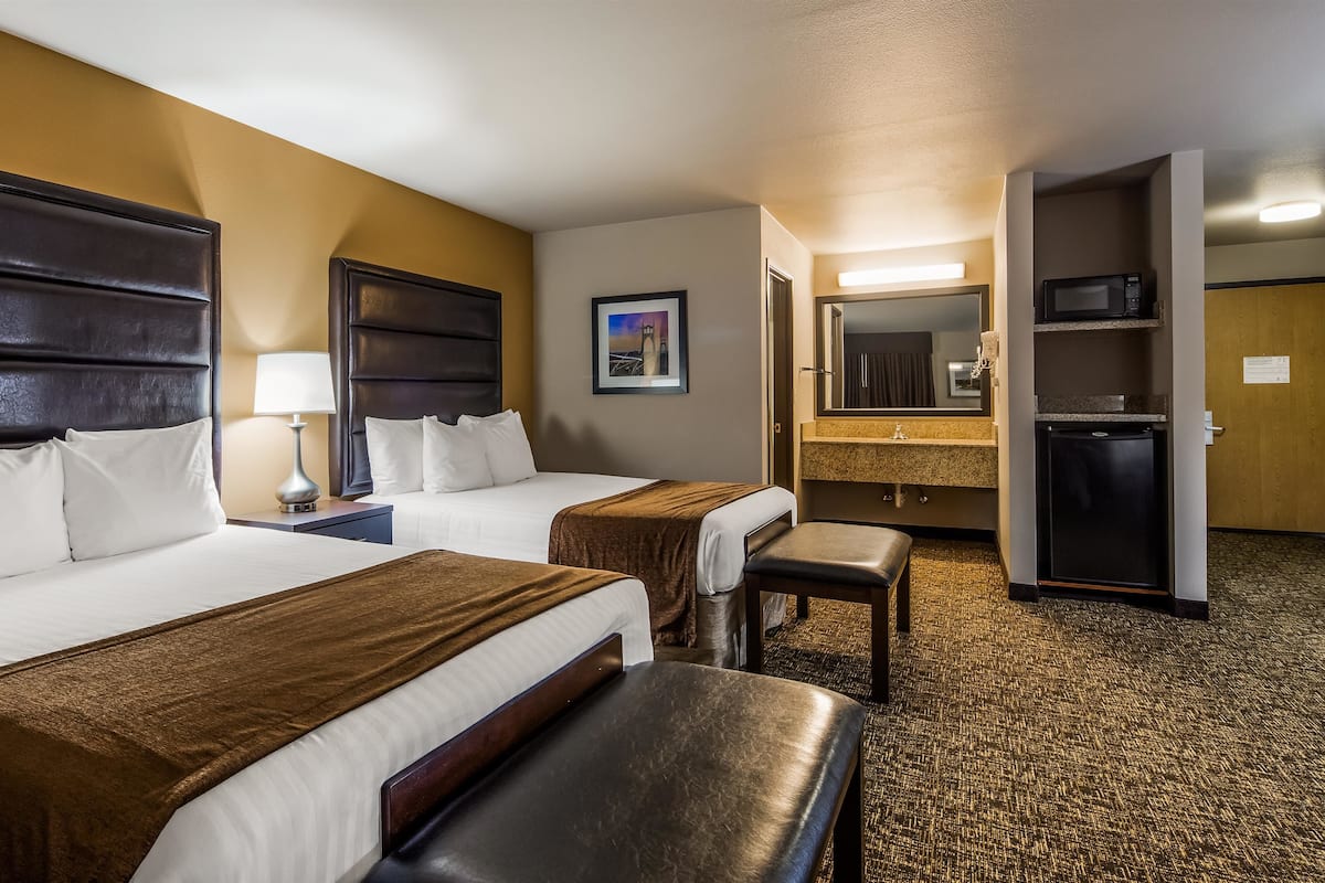 A hotel room with two beds, a bench, a desk, a lamp, a mirror, a microwave, and a small refrigerator.