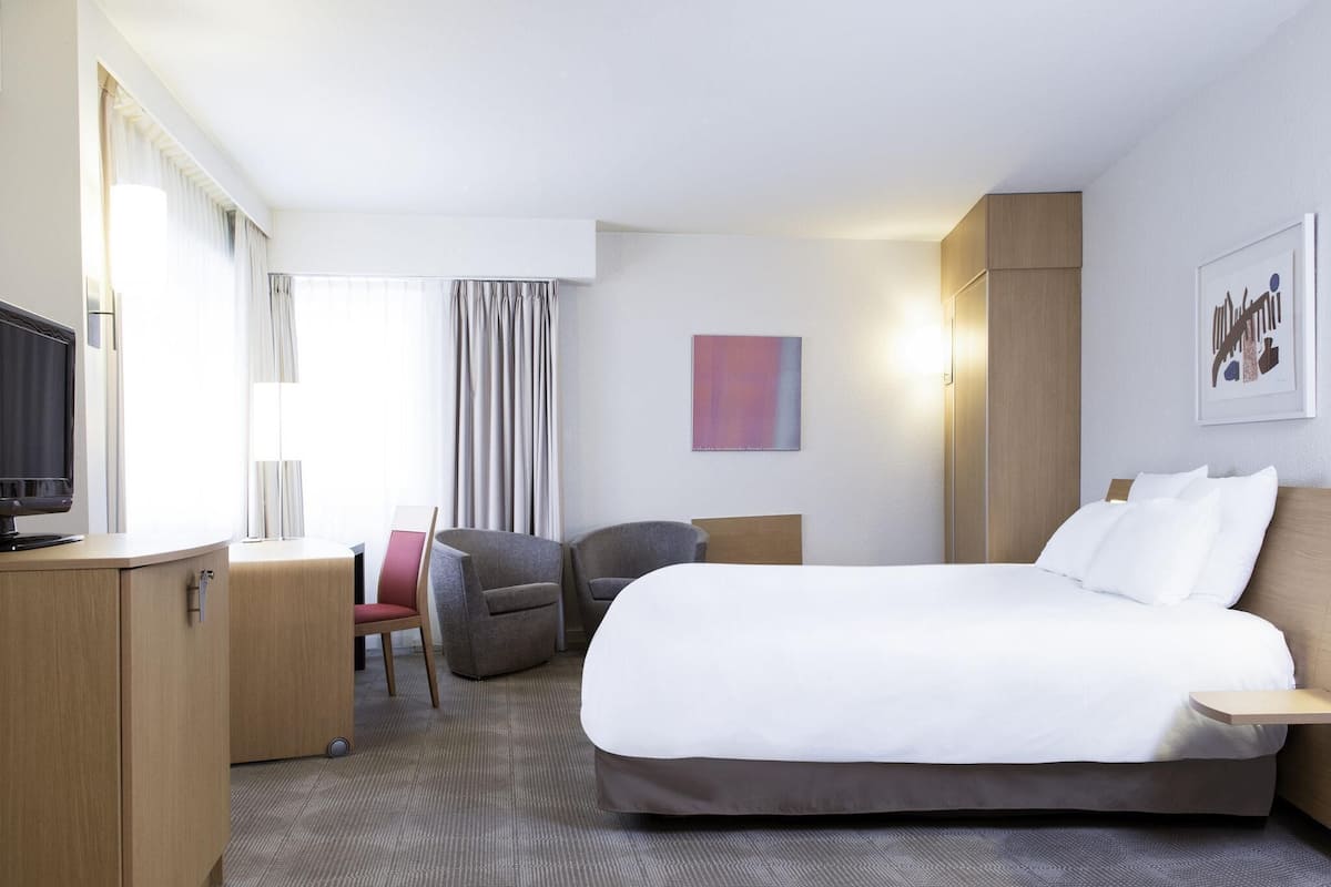 Ett hotellrum med en säng, ett skrivbord, en stol, en tv och ett fönster med gardiner.