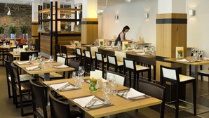 Un restaurant avec plusieurs tables dressées, nappées de serviettes blanches, avec couverts et verres à vin. Un serveur s’occupe d’une table.