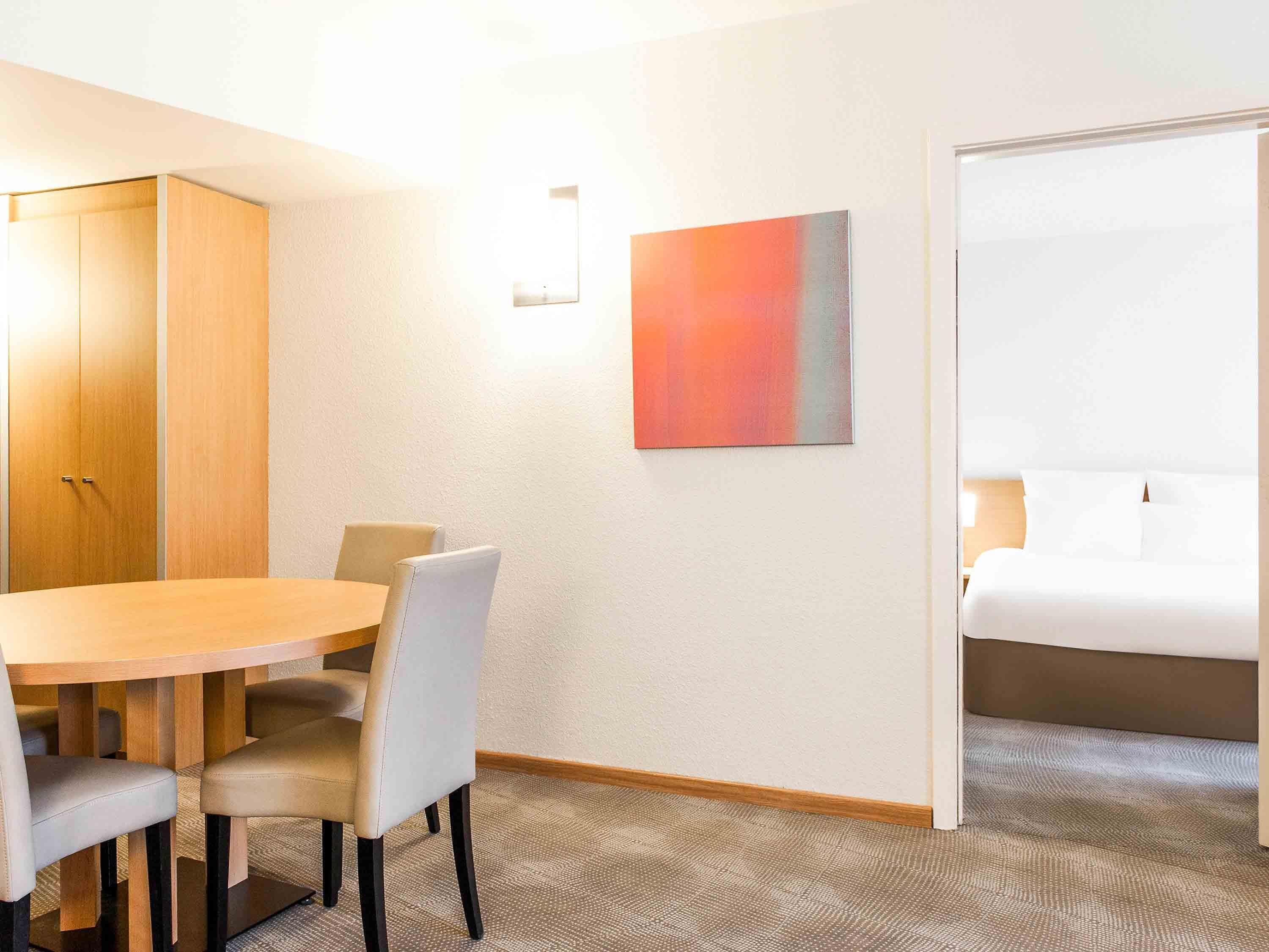 A hotel room with a wooden table, two chairs, a bed, and a painting on the wall.