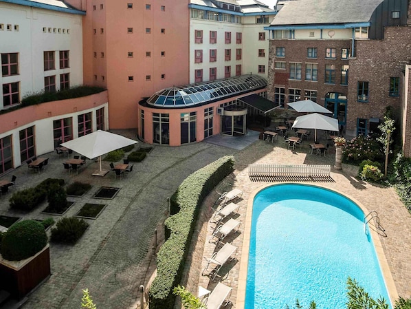 Un patio de hotel con piscina, mobiliario exterior y una zona cubierta con mesas y sombrillas.